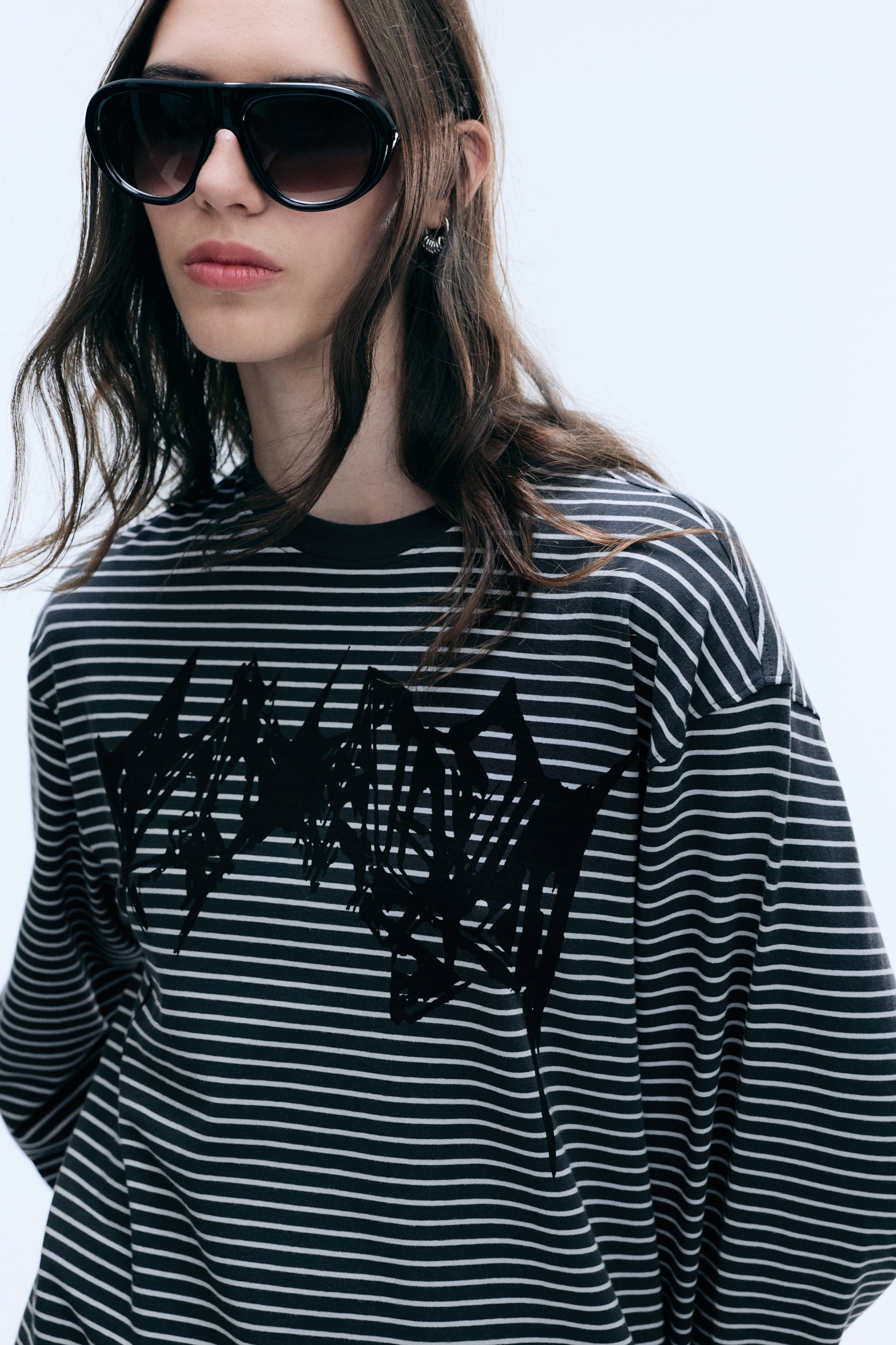 Grösseres Bild ansehen: Das Model trägt ein oversized Langarmshirt mit Grafik in einem schwarzen und grauen Querstreifenmuster. Das Shirt verfügt über einen Rundhalsausschnitt, überschnittene Schultern und einen grossen, abstrakten schwarzen Print auf der Brust.