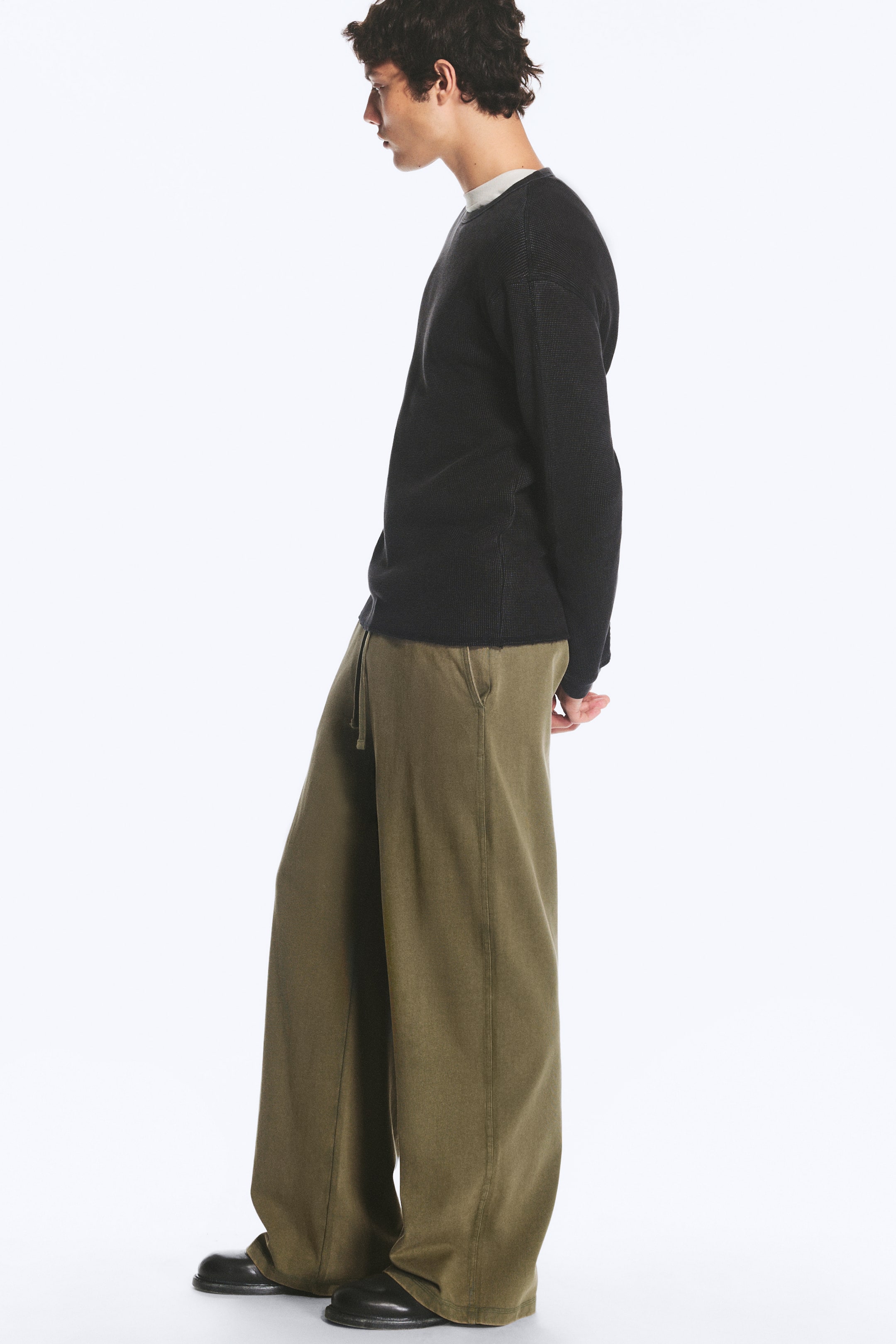Grotere afbeelding bekijken: De model draagt olijfgroene loungebroek met wijde pijpen, voorzien van een elastische tailleband met trekkoord en schuine zijzakken. De broek met relaxed fit valt over zwarte leren schoenen en is gecombineerd met een zwarte trui met lange mouwen.