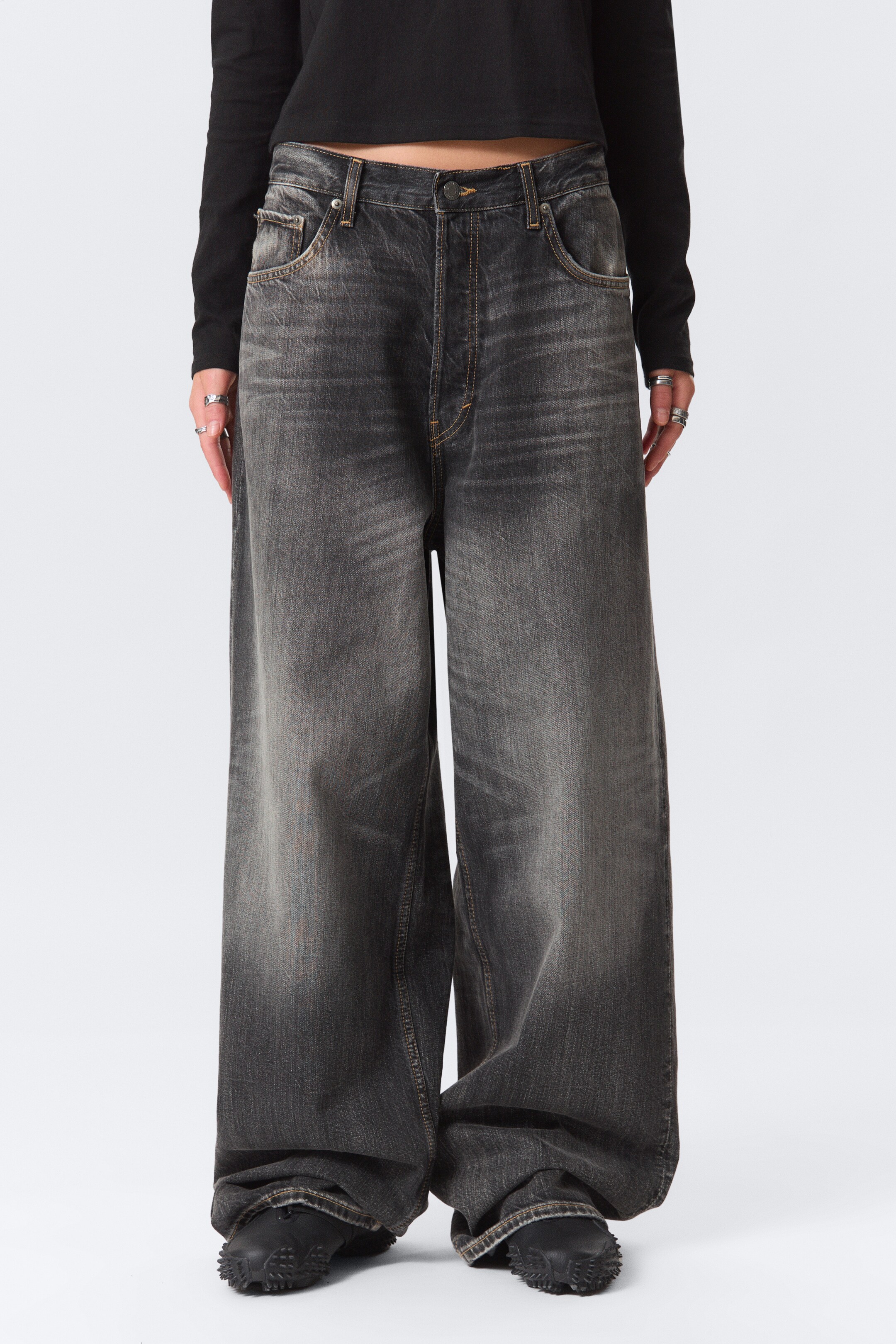 Grösseres Bild ansehen: Ein Paar dunkelgraue, weit geschnittene Jeans wird präsentiert, nach vorne gerichtet. Die Jeans zeichnen sich durch eine verblasste Waschung mit sichtbaren Falten, eine hohe Taille mit Knopfverschluss und kontrastierende hellbraune Nähte an den Nähten und vorderen Schaufeltaschen aus; ihre voluminösen Beine sammeln sich über schwarzem Schuhwerk.