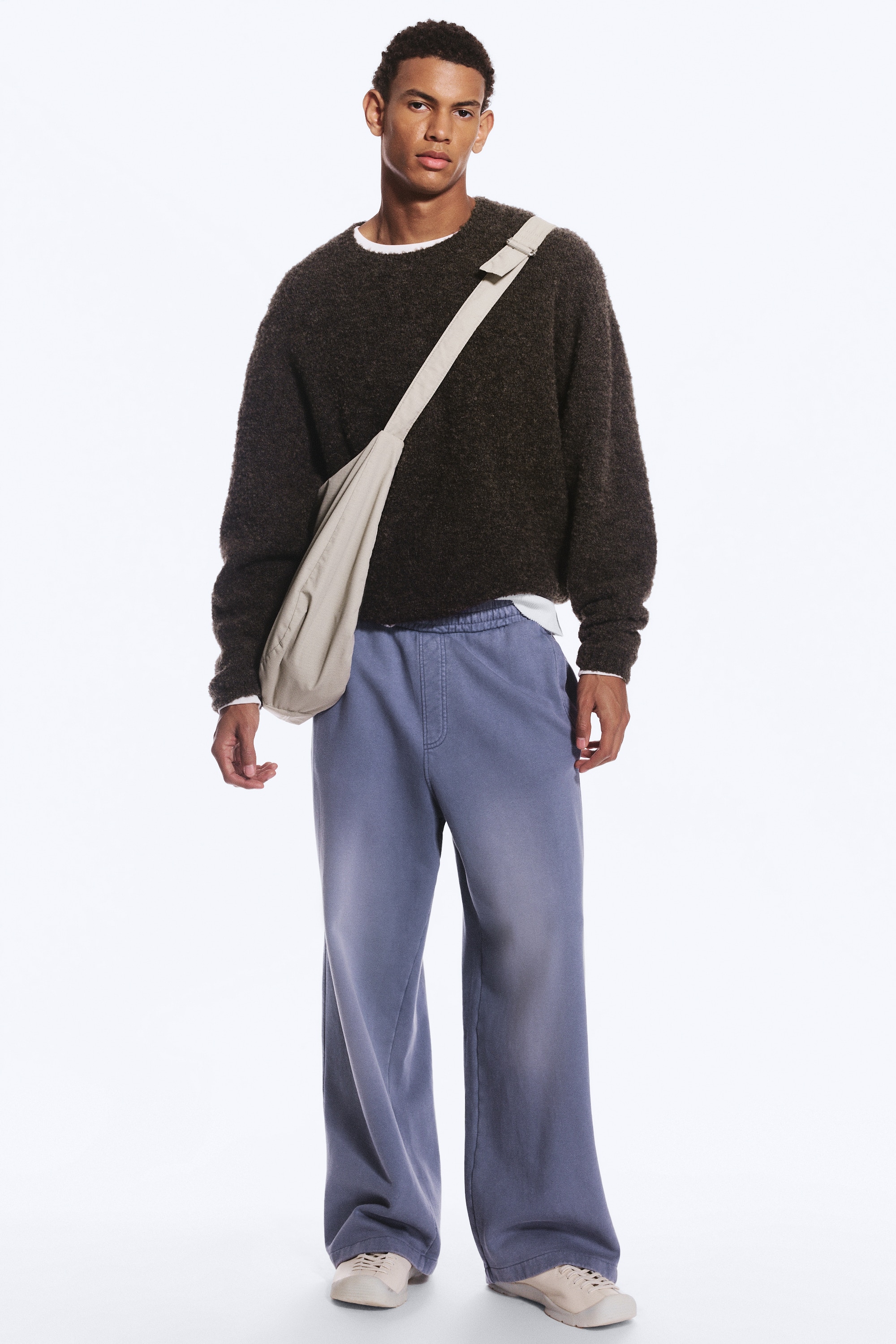 I pantaloni della tuta, visti frontalmente, presentano un colore blu-grigio sbiadito con una cintura elasticizzata e un dettaglio di patta finta. Mostrano tasche laterali a filetto, gambe larghe e dritte, e orli a taglio vivo.