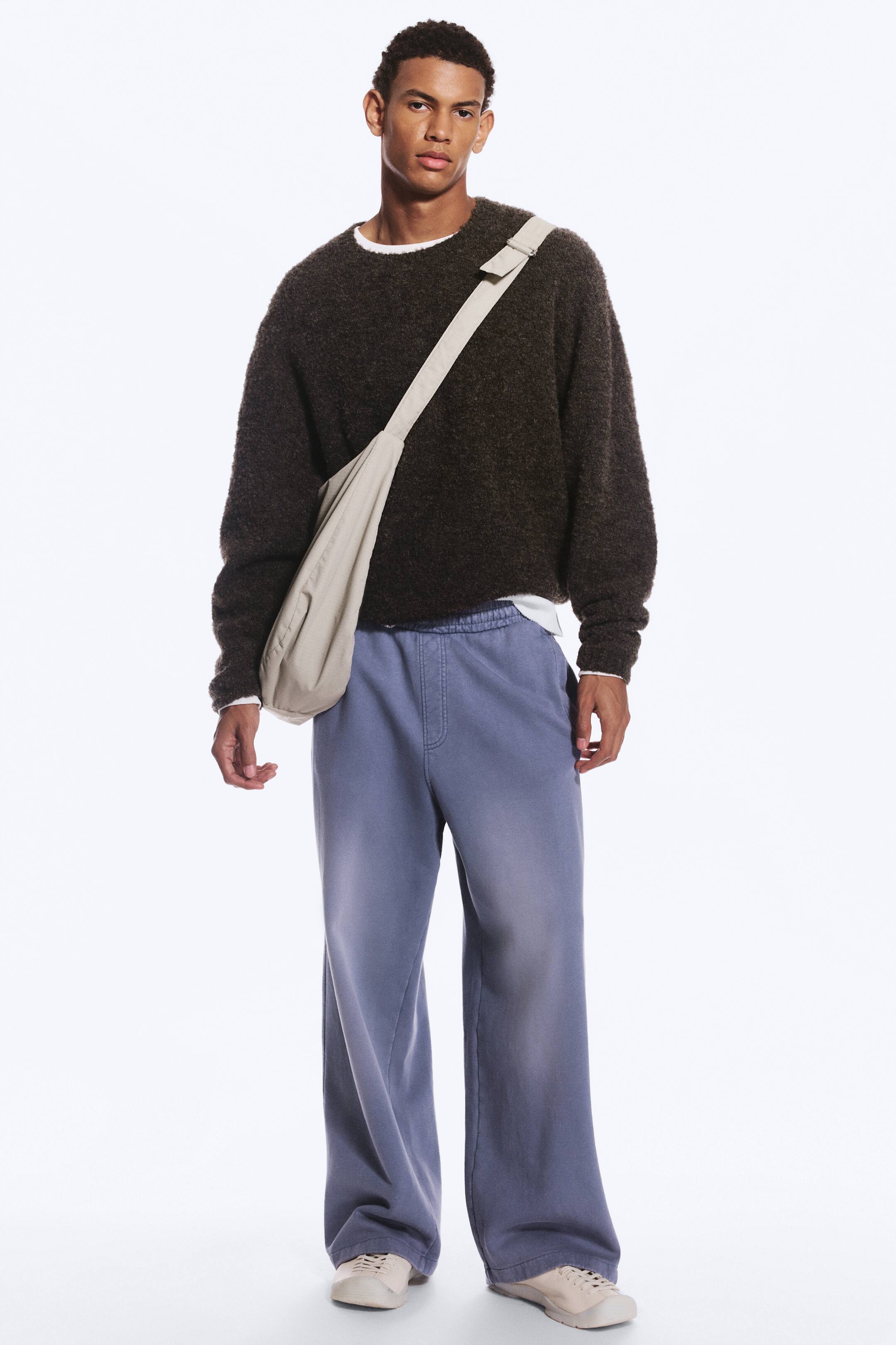 Grotere afbeelding bekijken: Een model draagt een losvallende, vervaagde blauwe sweatpants met een elastische tailleband met middelhoge taille en een nep gulpdetail. De broek met wijde pijpen wordt gecombineerd met een donkerbruine sweater met textuur, een lichtgekleurde crossbodytas en beige sneakers.