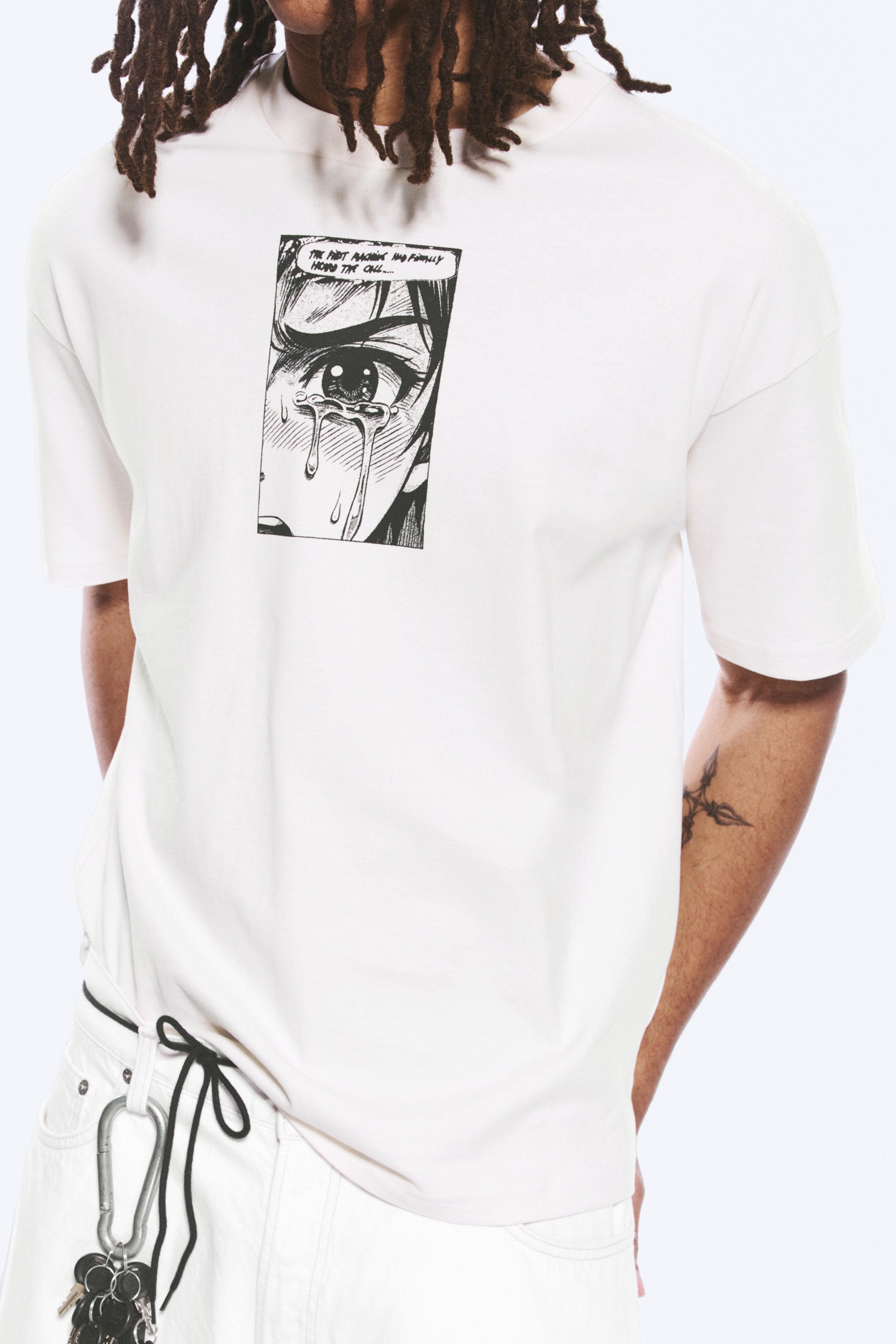Se større billede: En hvid T-shirt bæres, der har en løs pasform, en høj rund hals og korte ærmer. Et sort-hvidt manga-stil tegneseriepanel, der viser et grædende øje med teksten "THE FIRST ANCHOR HAS FINALLY HEARD THE CALL...", er trykt på forsiden.