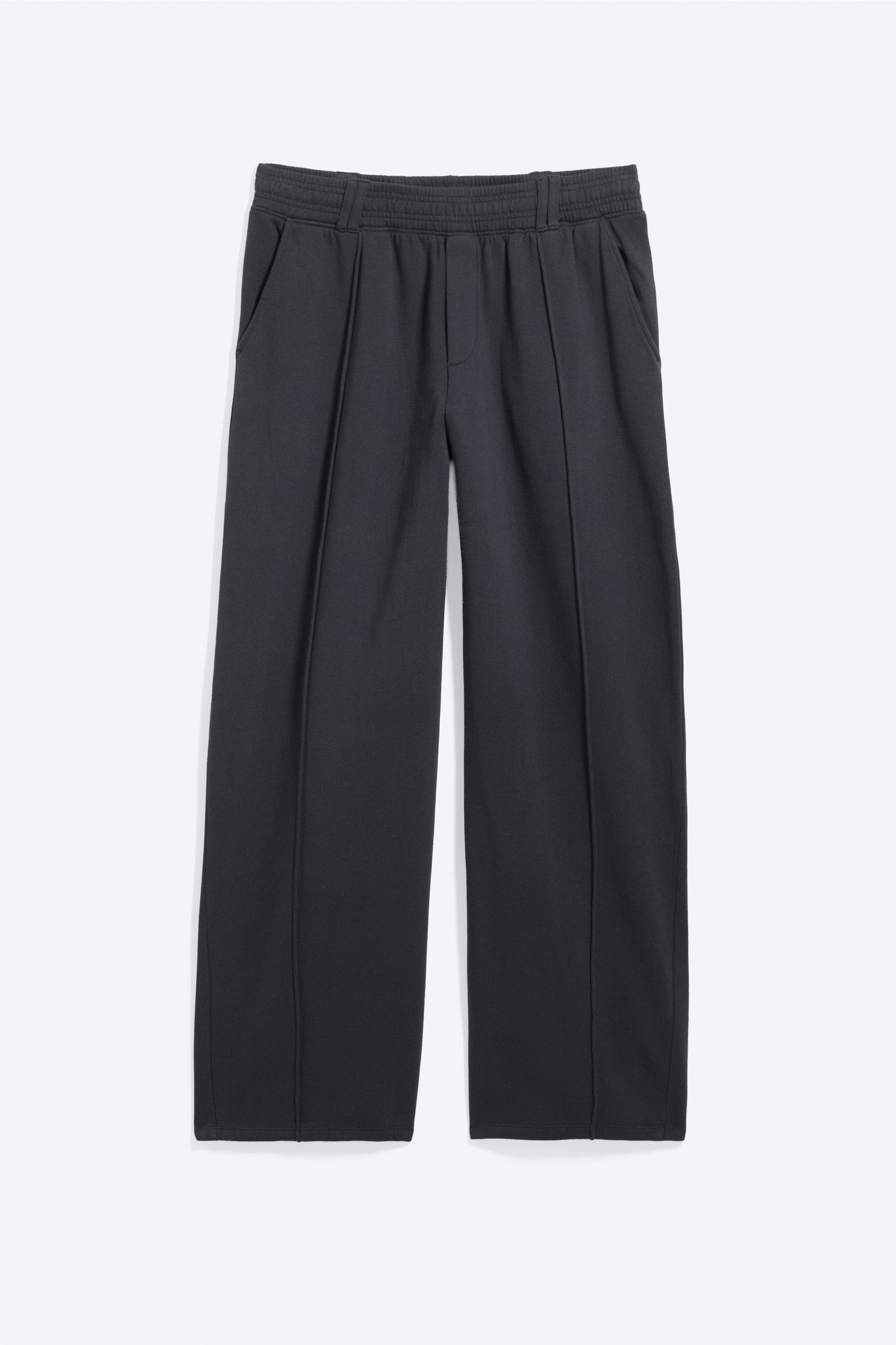 De losse geplooide joggers in donkergrijs worden gedragen door het model en hebben een ontspannen, wijduitlopende pasvorm met duidelijke middennaden die langs de voorkant van elk broekspijp lopen. De joggers zijn gestyled met een zwarte riem door de riemlussen van de tailleband, een cropped sweatshirt met lange mouwen en zwarte sneakers.