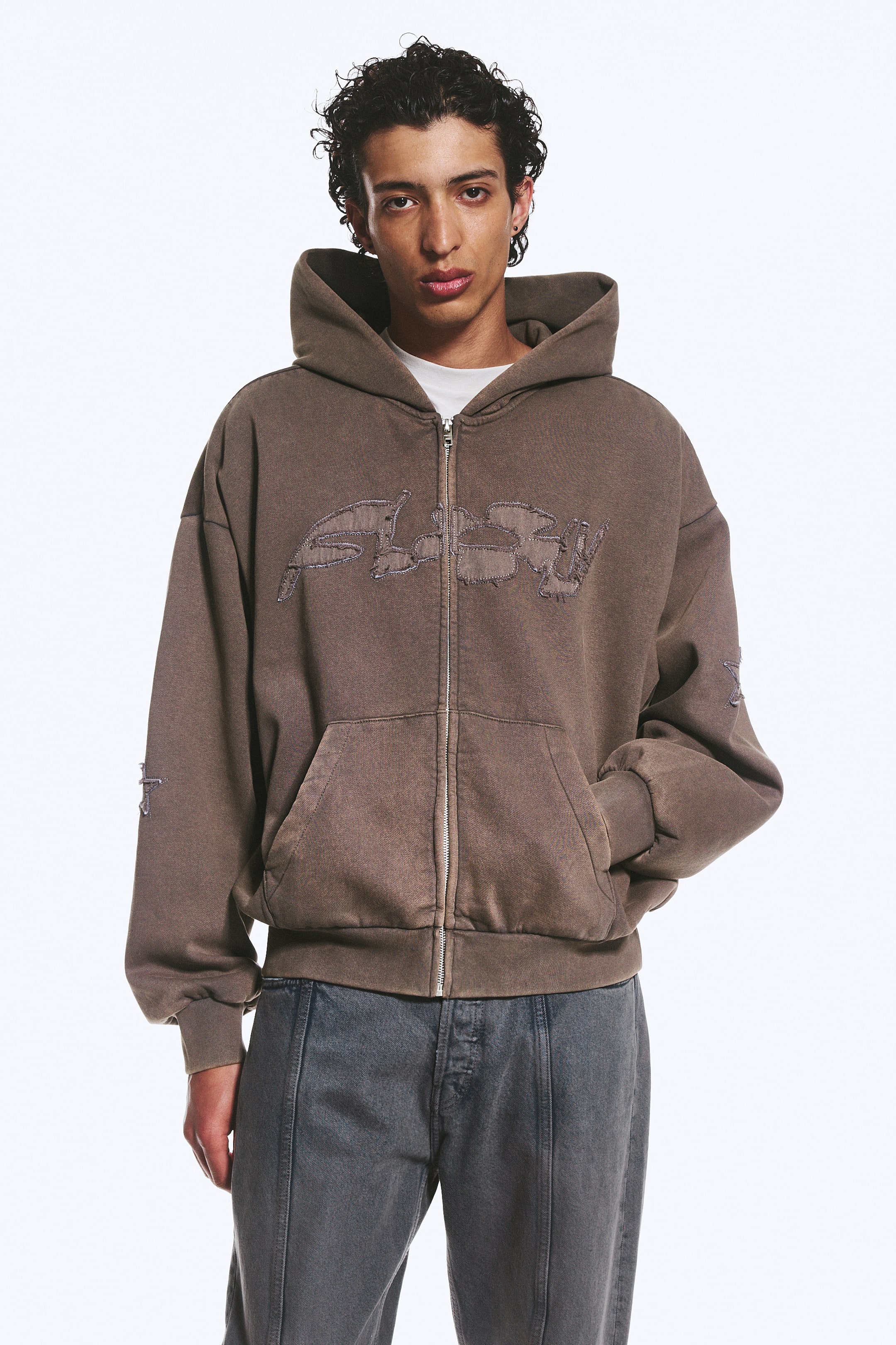 View larger image: A washed brown zip-up hoodie is facing forward, featuring a full-zip fastening, an attached hood, and a large distressed embroidered text design across the chest. It also showcases front pockets, ribbed cuffs and hem, and a small embroidered star on the left sleeve, with a white t-shirt visible underneath.