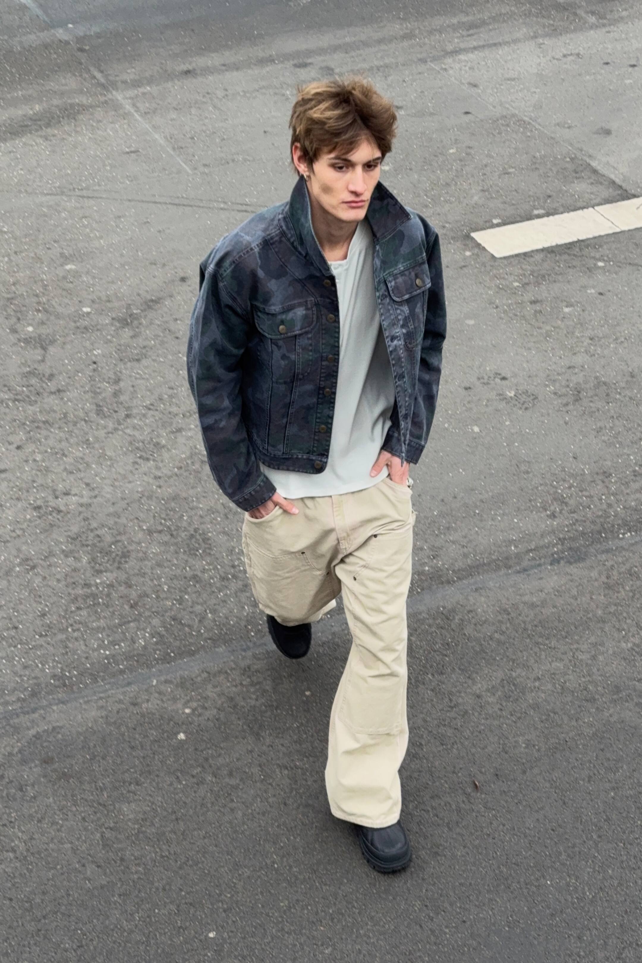 Se større billede: En mørk camouflage-mønstret denimjakke bæres åbent over en lys t-shirt. Den har en nedfoldet krave, to knappede brystlommer, to sidelommer og knappede manchetter.