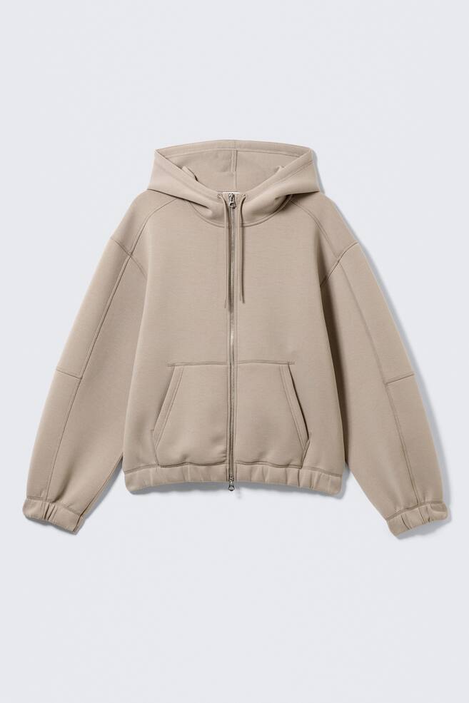 Ein beige Kapuzenpullover mit durchgehendem Reißverschluss präsentiert seine VORDERSEITE, ausgestattet mit einem silbernen Reißverschluss, einer Kordelkapuze, einer geteilten Kängurutasche, sichtbaren Teilungsnähten an den Ärmeln und Schultern sowie gerippten Bündchen und Saum. - 1