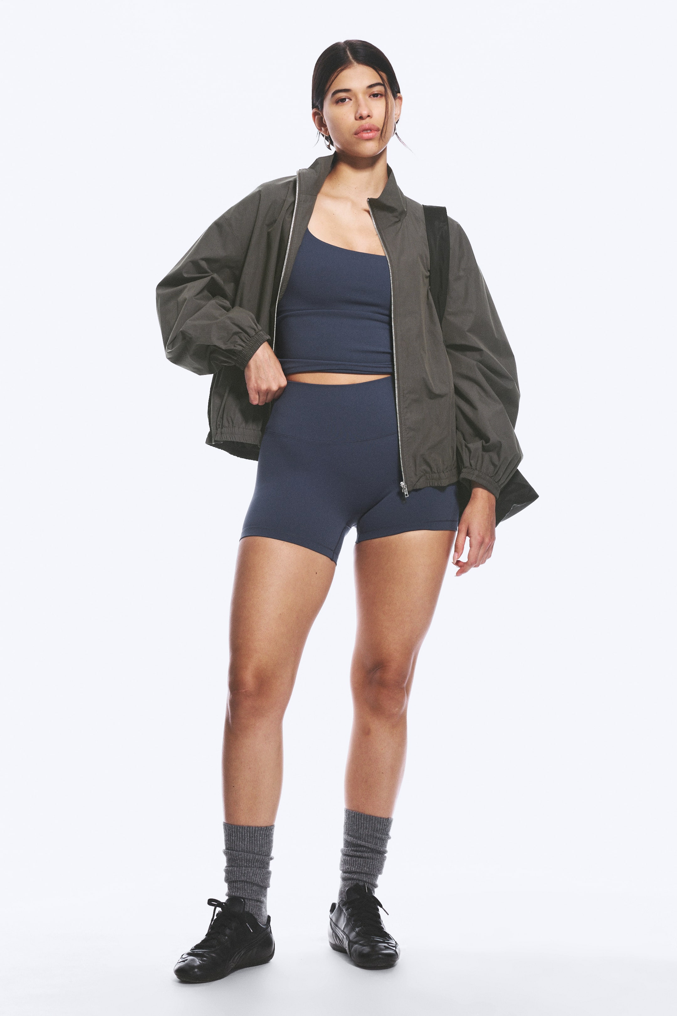 Visualizza immagine più grande: Un bomber verde oliva scuro è indossato aperto, rivelando un top corto blu navy con scollo a barchetta e pantaloncini sportivi a vita alta blu navy. La giacca presenta una chiusura frontale con zip completa e polsini elasticizzati, mentre calzini grigi a coste e scarpe sportive nere completano l'ensemble.