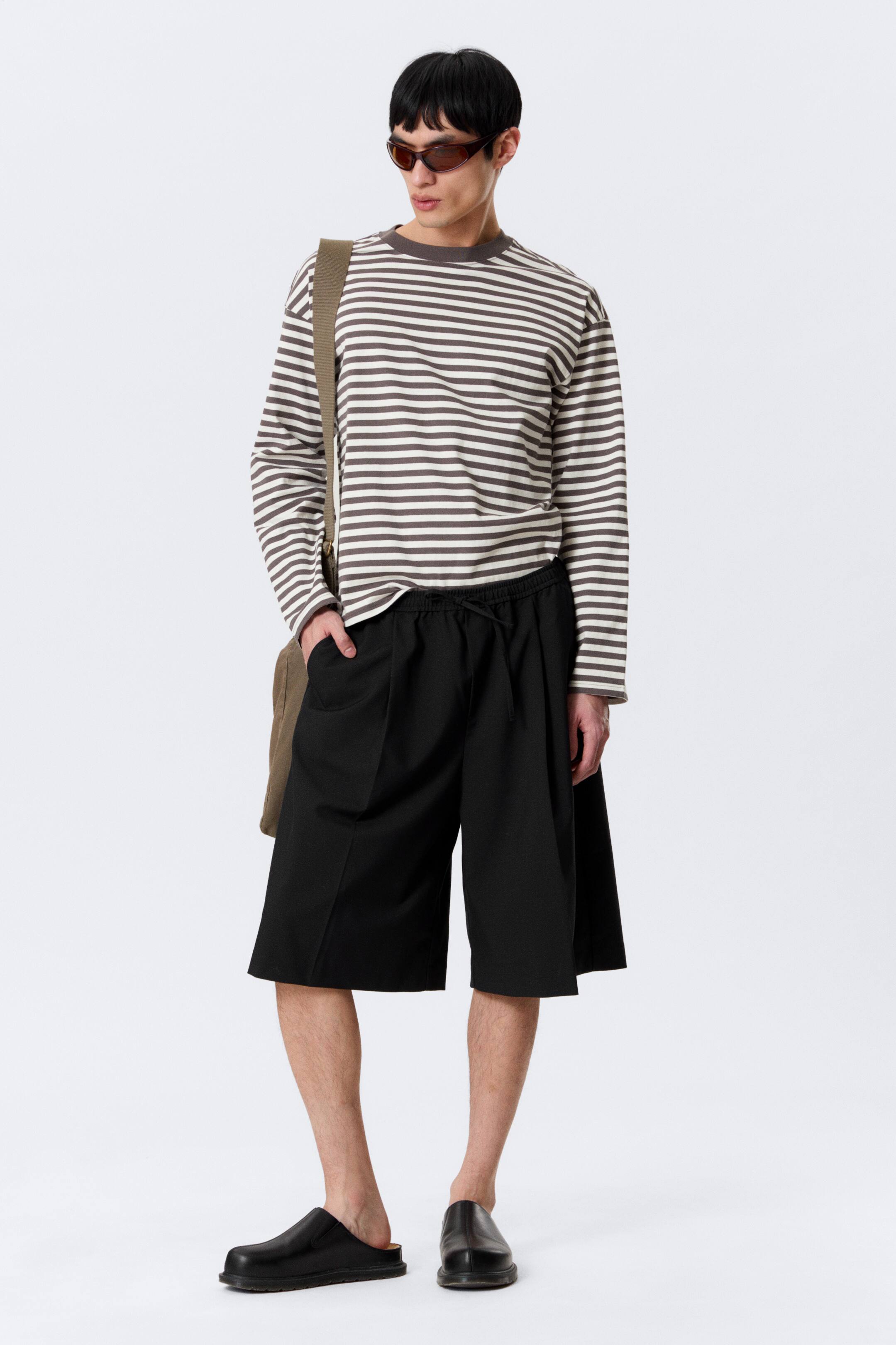 Visualizza immagine più grande: Una maglietta a maniche lunghe, caratterizzata da righe orizzontali color crema e marrone con girocollo e polsini a coste, viene presentata con pantaloncini neri a gamba larga che arrivano sotto il ginocchio e presentano una vita elastica con coulisse e pieghe frontali. Scarpe nere senza lacci completano l'insieme.