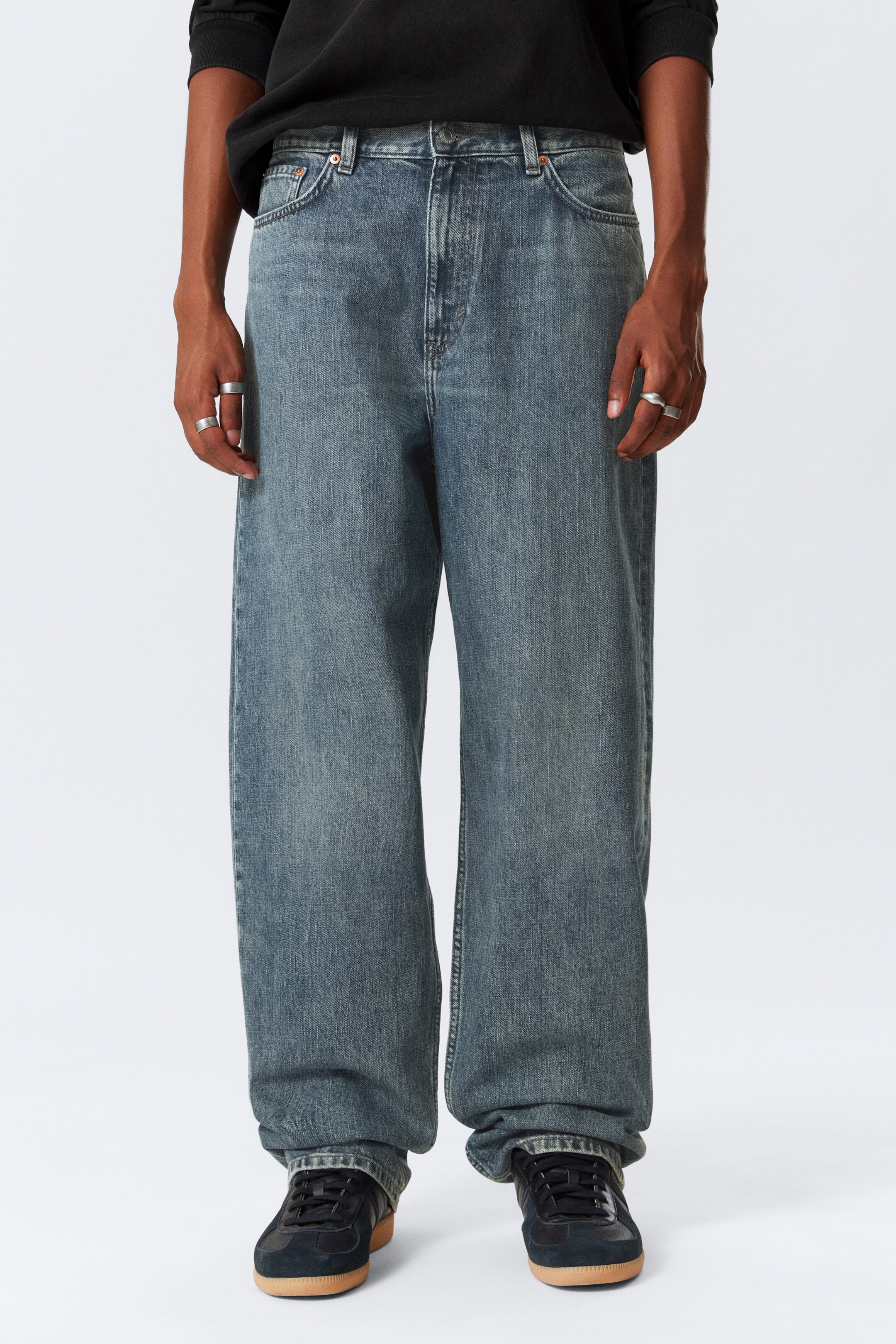 Größeres Bild anzeigen: Gewaschene blaue, weit geschnittene Jeans werden frontal getragen, mit einer lockeren Passform, einem Knopfverschluss und klassischem Fünf-Taschen-Design mit sichtbaren Kontrastnähten und Nieten. Ihr langer Saum sammelt sich über dunklen Sneakers.