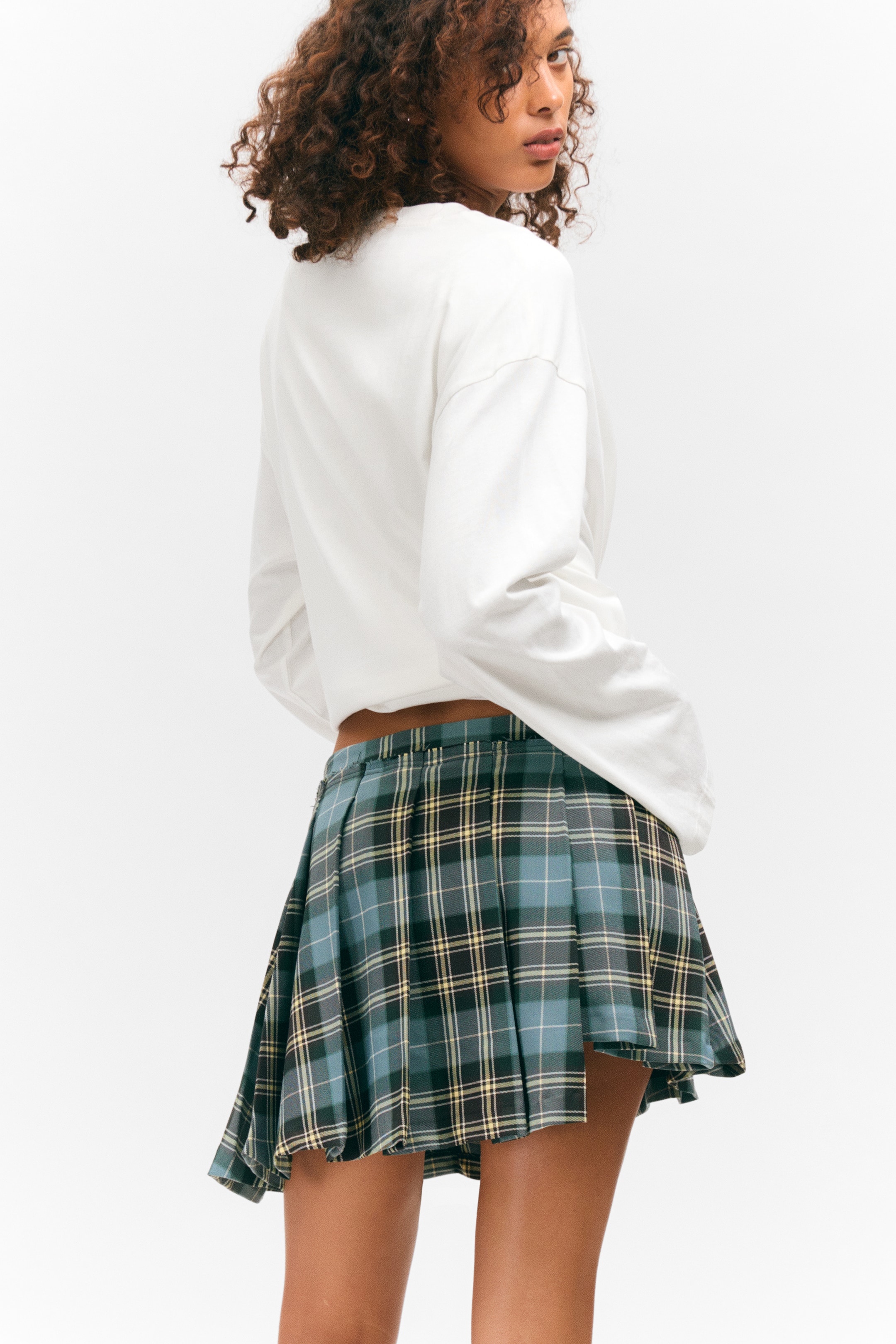 View larger image: A plaid pleated mini skirt is being presented from a back-side angle, featuring a blue, green, yellow, and black plaid pattern with numerous small pleats creating a flared design. A white long-sleeve top is partially visible, ending just above the skirt's waistband.
