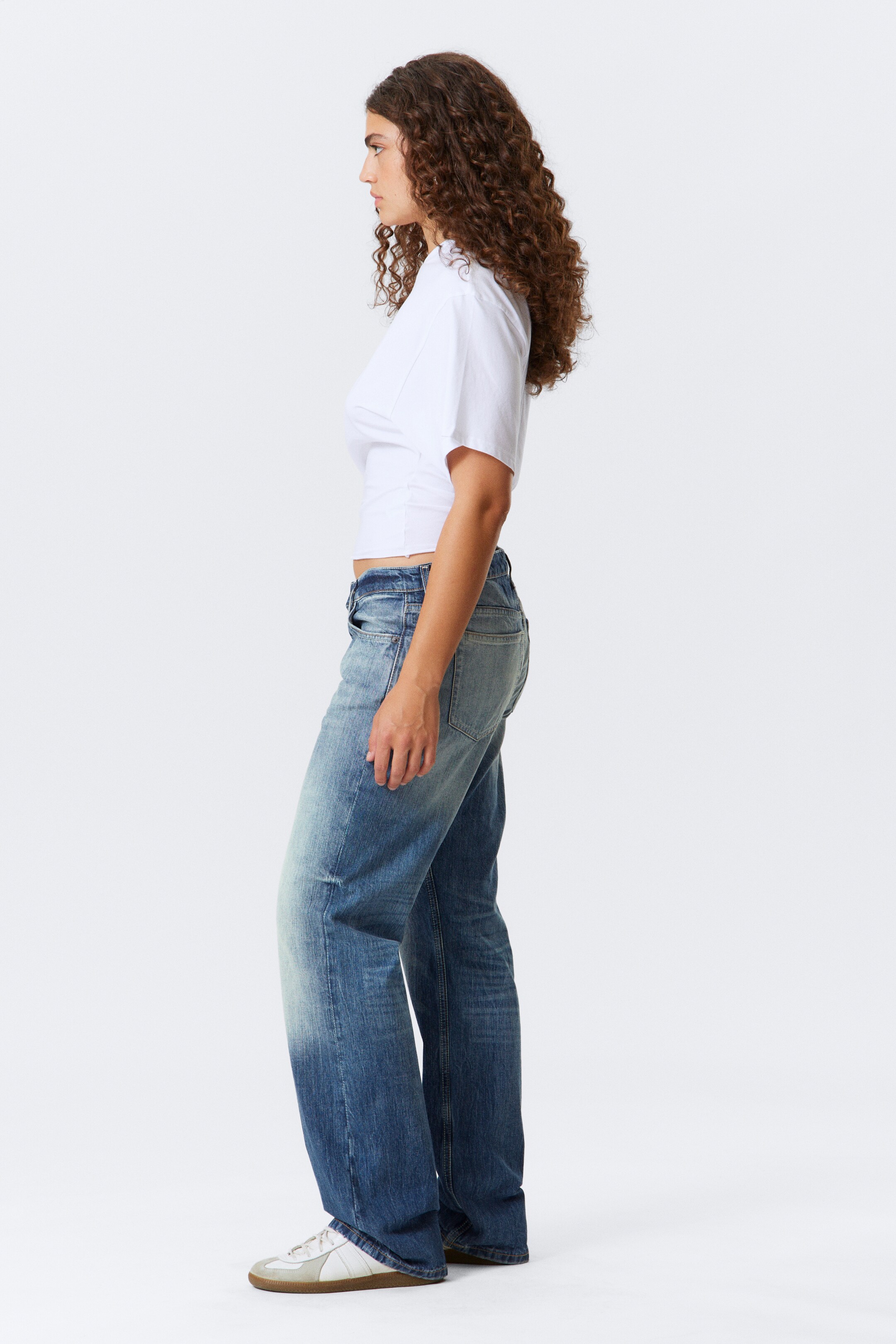 Agrandir l'image: Le produit, vu de côté, est un t-shirt court à manches courtes blanc avec un ourlet brut et une taille légèrement cintrée, associé à un jean bleu taille haute à jambes larges avec un effet délavé. Des baskets basses blanches et beiges sont également portées.