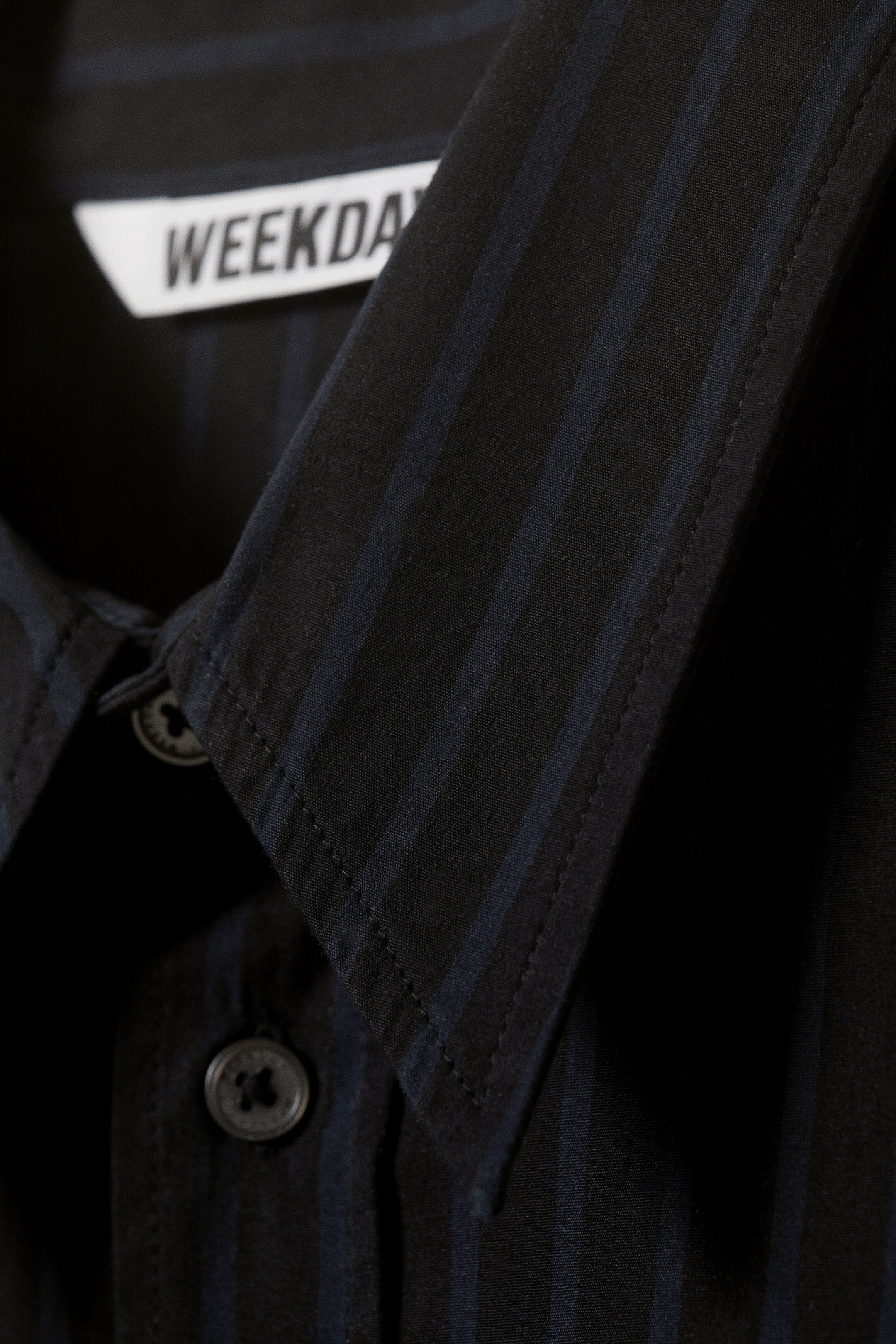 Ver imagen más grande: Una camisa oscura con sutiles rayas verticales se muestra en un primer plano, destacando su cuello y la tapeta frontal con botones oscuros. Una etiqueta de marca blanca es visible en el interior del cuello.