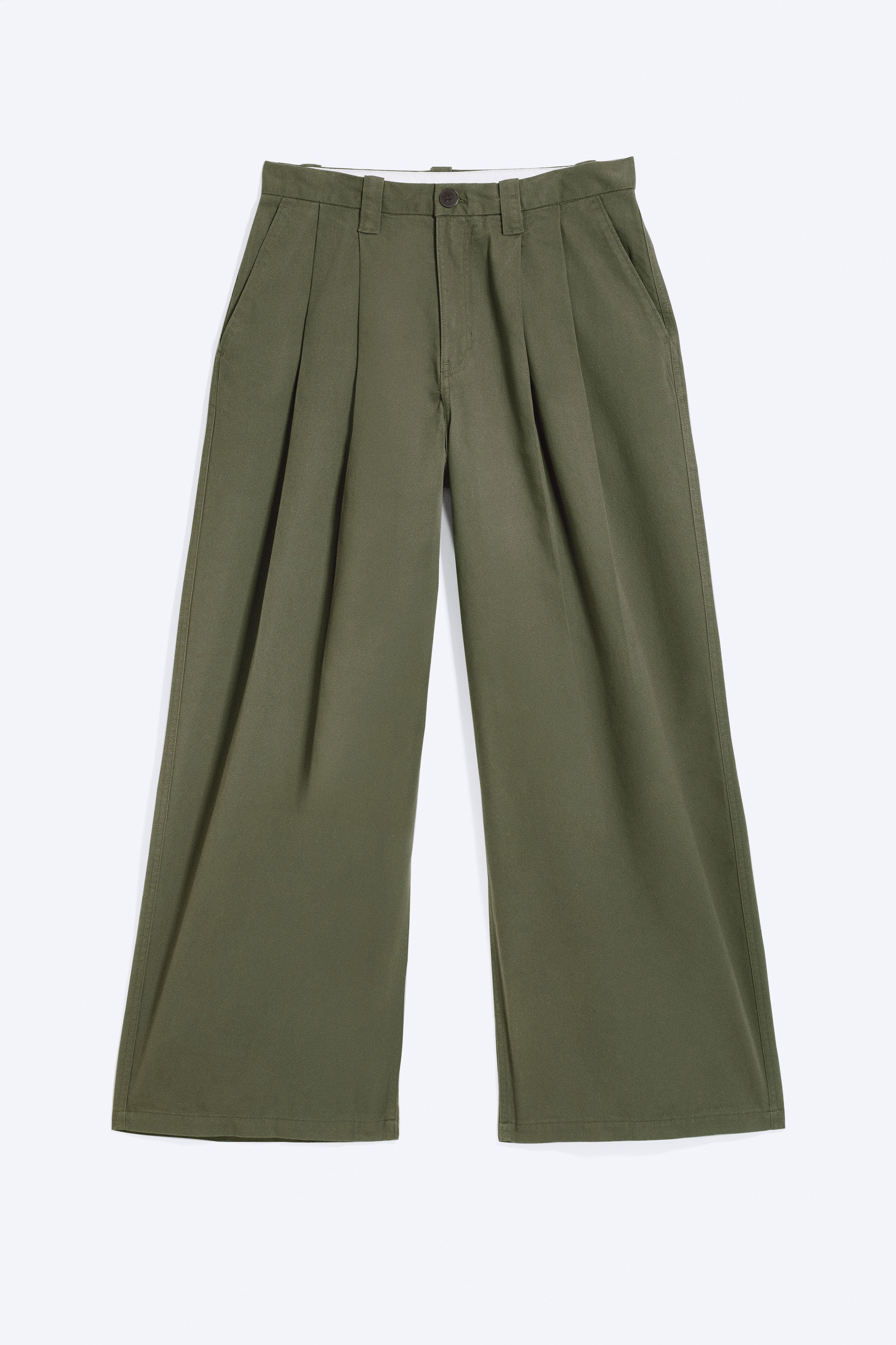 Loose-fit chino trousers in olive green are being worn by the model, who is standing in profile with one hand resting in a slanted side pocket. The wide-leg trousers feature a baggy silhouette with front pleats and a back welt pocket, and are styled with a black knitted jumper and black shoes.