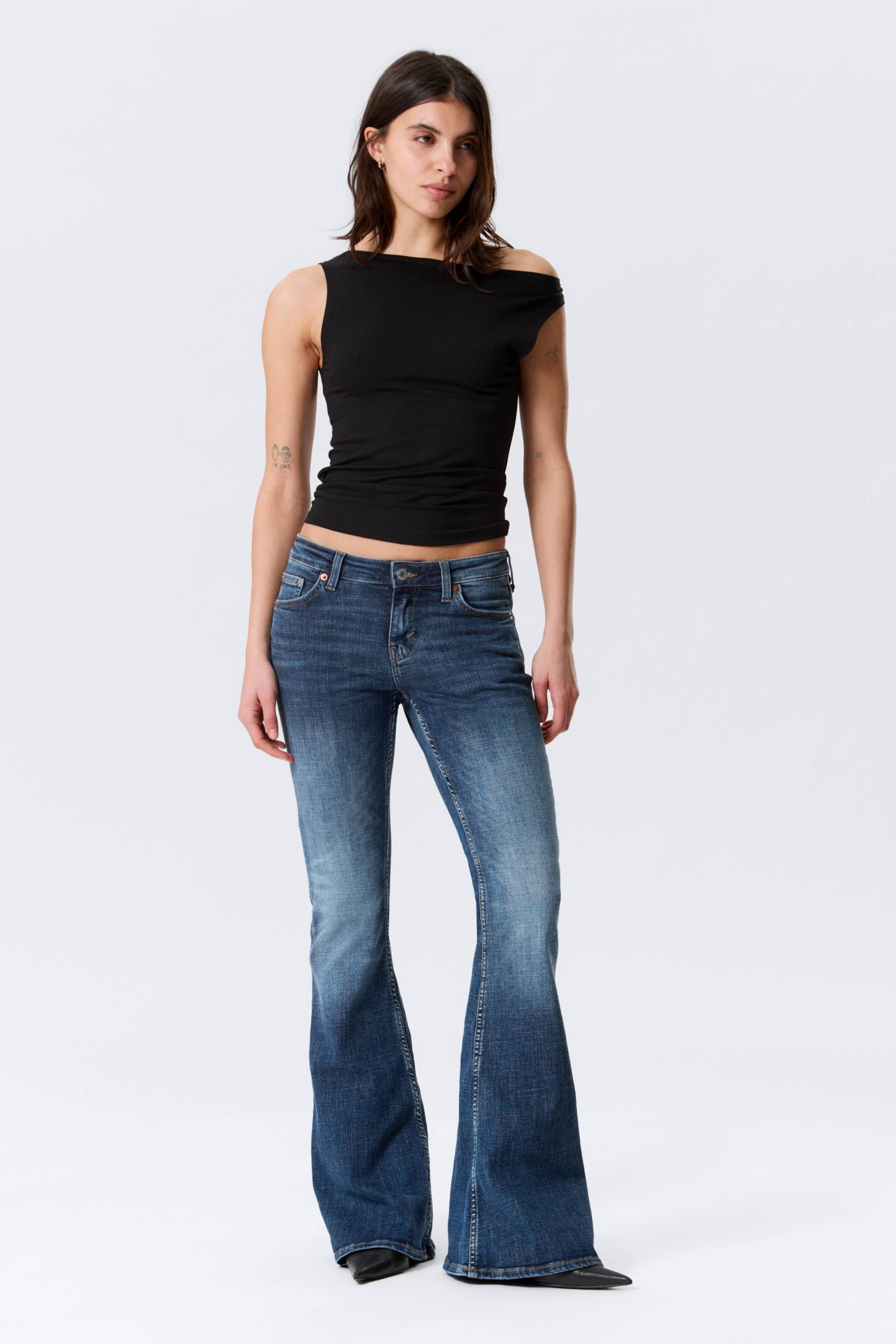 Visualizza immagine più grande: Un top nero senza maniche, caratterizzato da uno scollo asimmetrico con una spalla scoperta, è abbinato a jeans svasati in denim blu. I jeans presentano un lavaggio scolorito, una vita bassa con chiusura a bottoni e tasche frontali, e un'apertura svasata della gamba dal ginocchio.