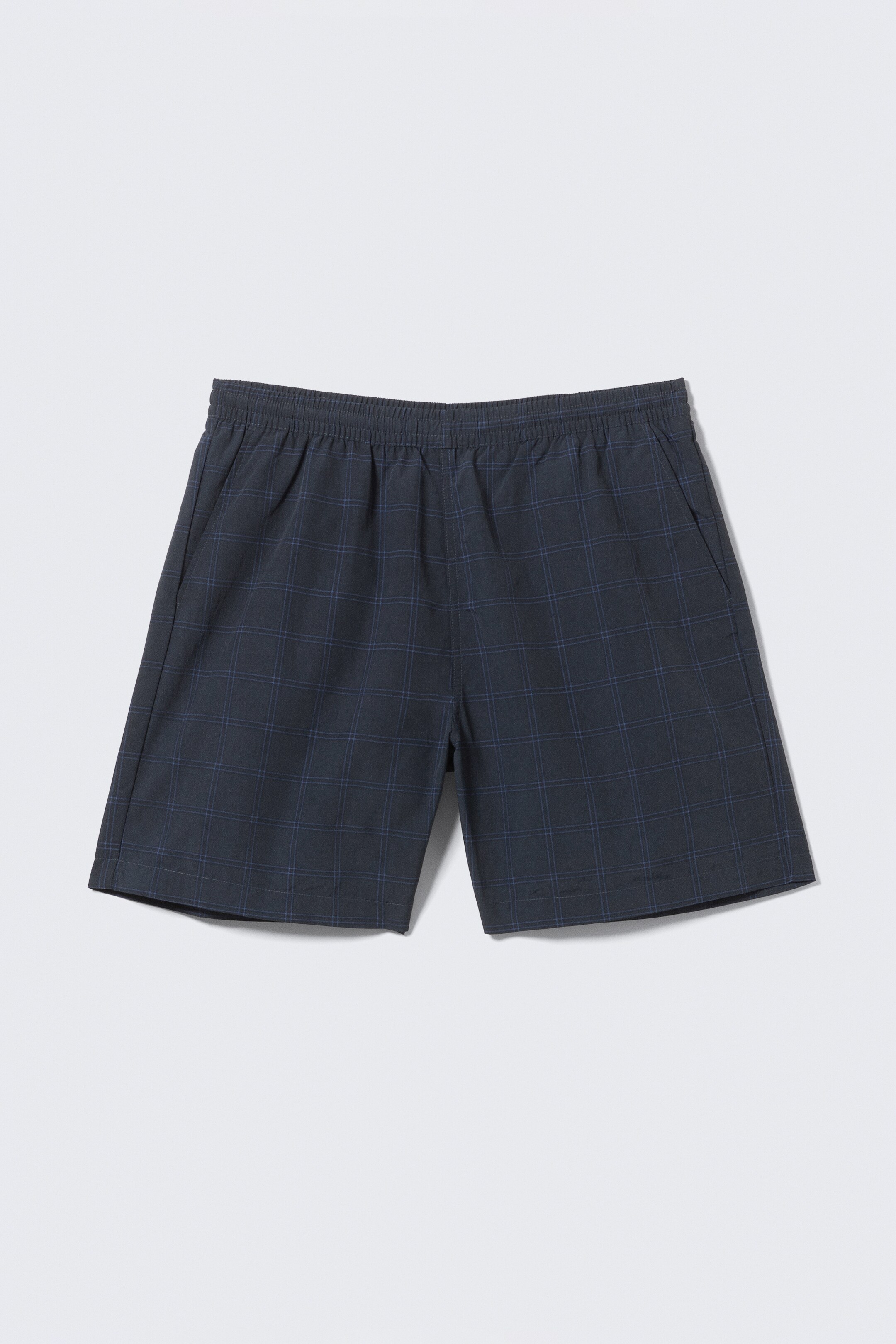 Zobrazit větší obrázek: Tmavě šedé kostkované plavky se subtilním modrým vzorem mřížky jsou zobrazeny zepředu. Mají boční kapsy s rozparkem a elastický pas.