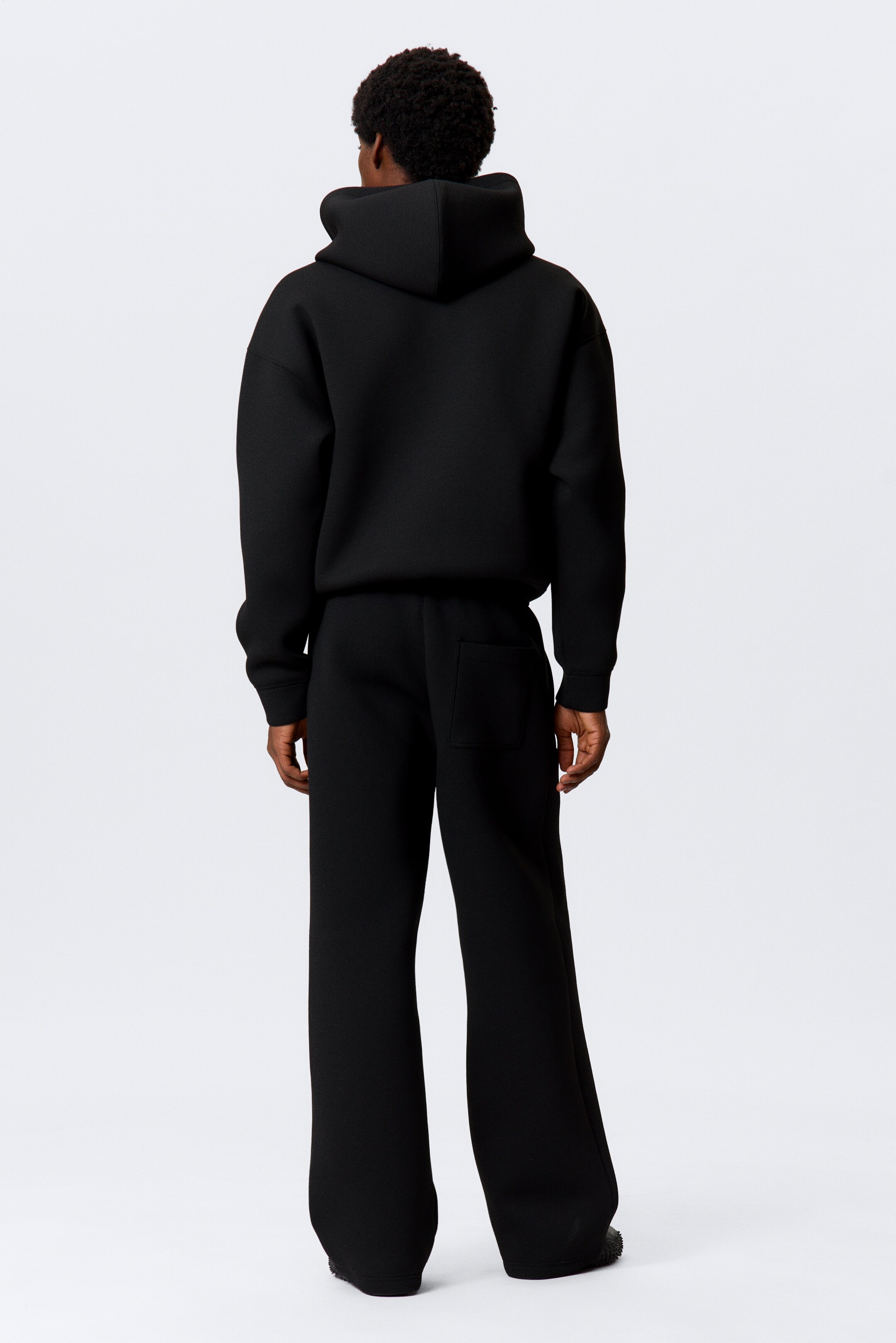 Agrandir l'image: Les pantalons de survêtement noirs à coupe ample, fabriqués en tissu scuba épais et présentant une silhouette décontractée à jambes larges avec une poche arrière visible, sont portés par le mannequin.