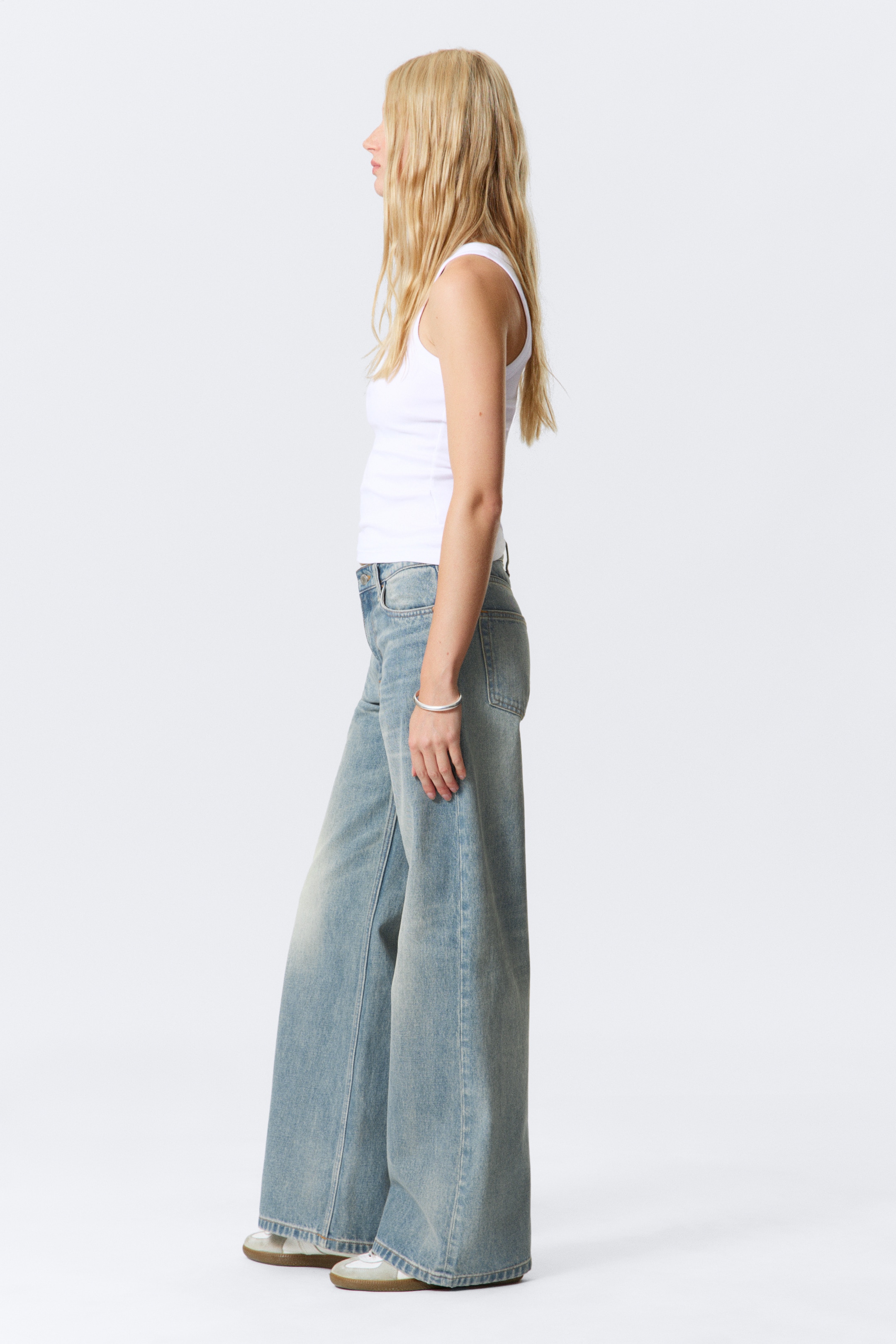 Visualizza immagine più grande: Jeans a vita media e gamba larga in un lavaggio chiaro sono indossati dalla modella, che è in piedi di profilo. I jeans presentano un design a cinque tasche e sono abbinati a un top bianco e sneakers di colore chiaro.