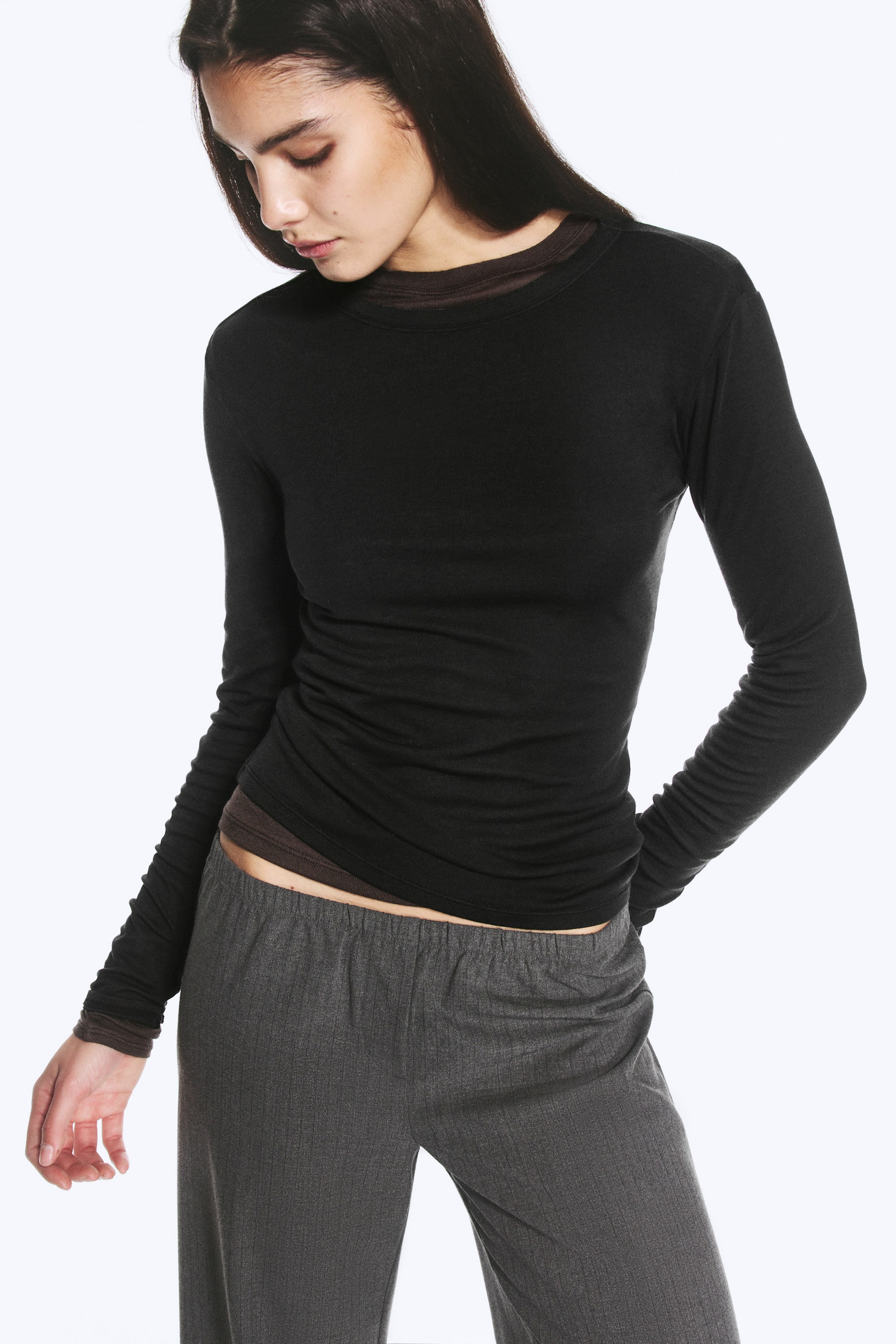 Agrandir l'image: Le modèle porte un haut en maille noir, ajusté et à manches longues, mettant en valeur sa coupe près du corps et son large col rond. Le haut est porté par-dessus une sous-couche marron à manches longues, visible au niveau du col, des poignets et de l’ourlet, et il est associé à un pantalon gris.
