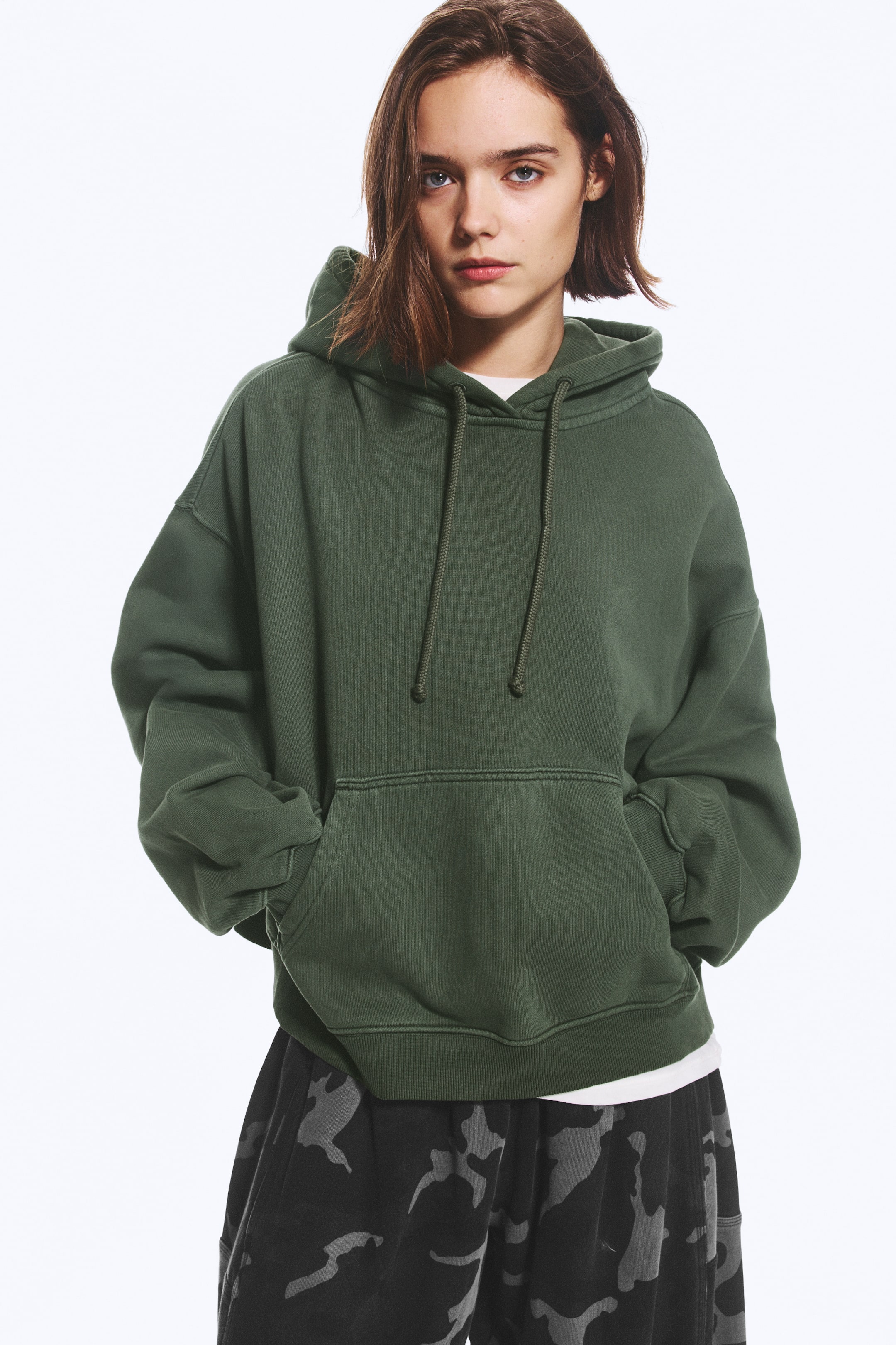 Visualizza l'immagine ingrandita: Una felpa con cappuccio verde oversize, con cappuccio con coulisse, spalle scese e orlo a costine, indossata dalla modella. La modella è in piedi con le mani infilate nella tasca a marsupio frontale.