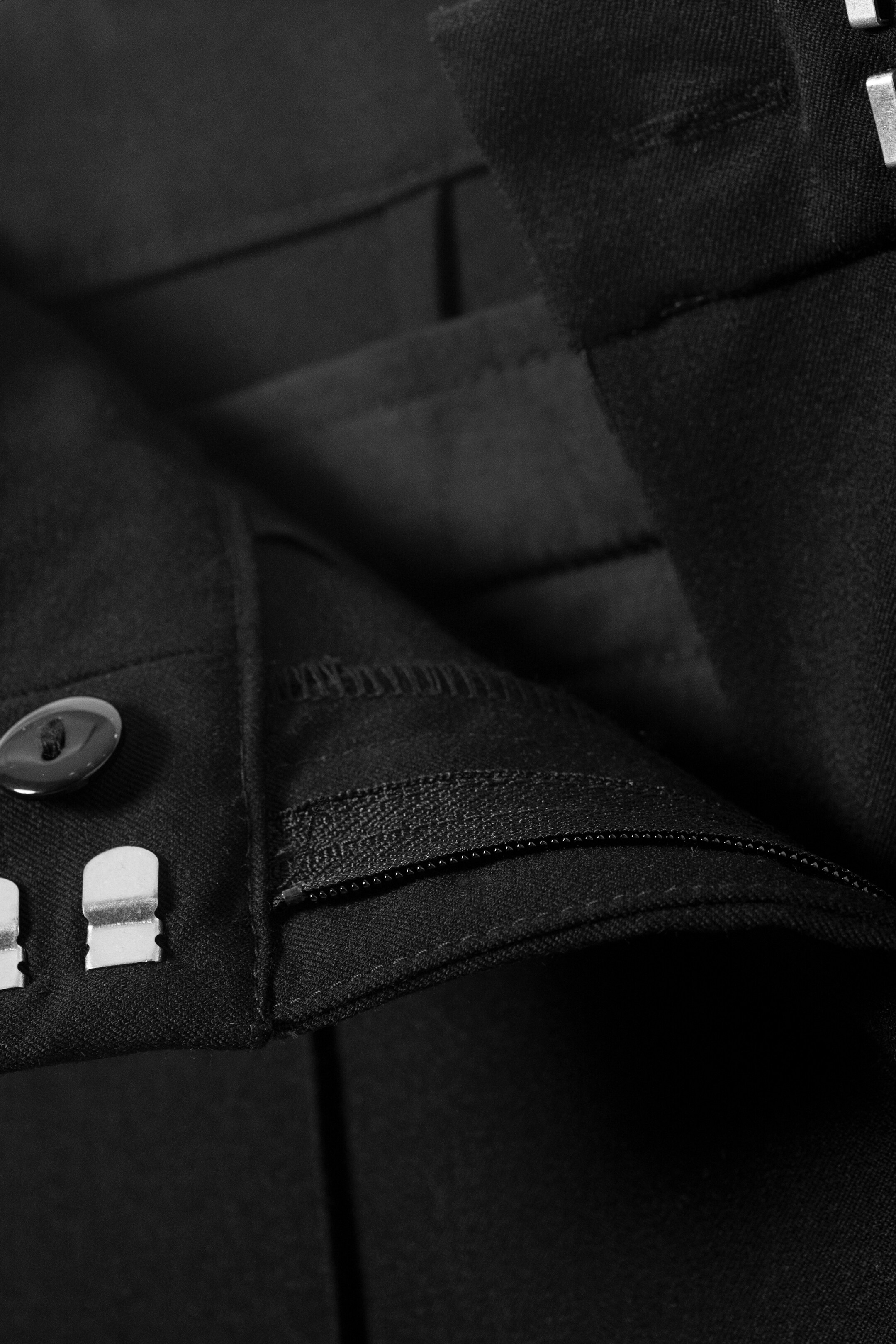View larger image: The front of black trousers is showing an open zippered fly, revealing the zipper. A button and two metal hook-and-bar closures are visible on the waistband.