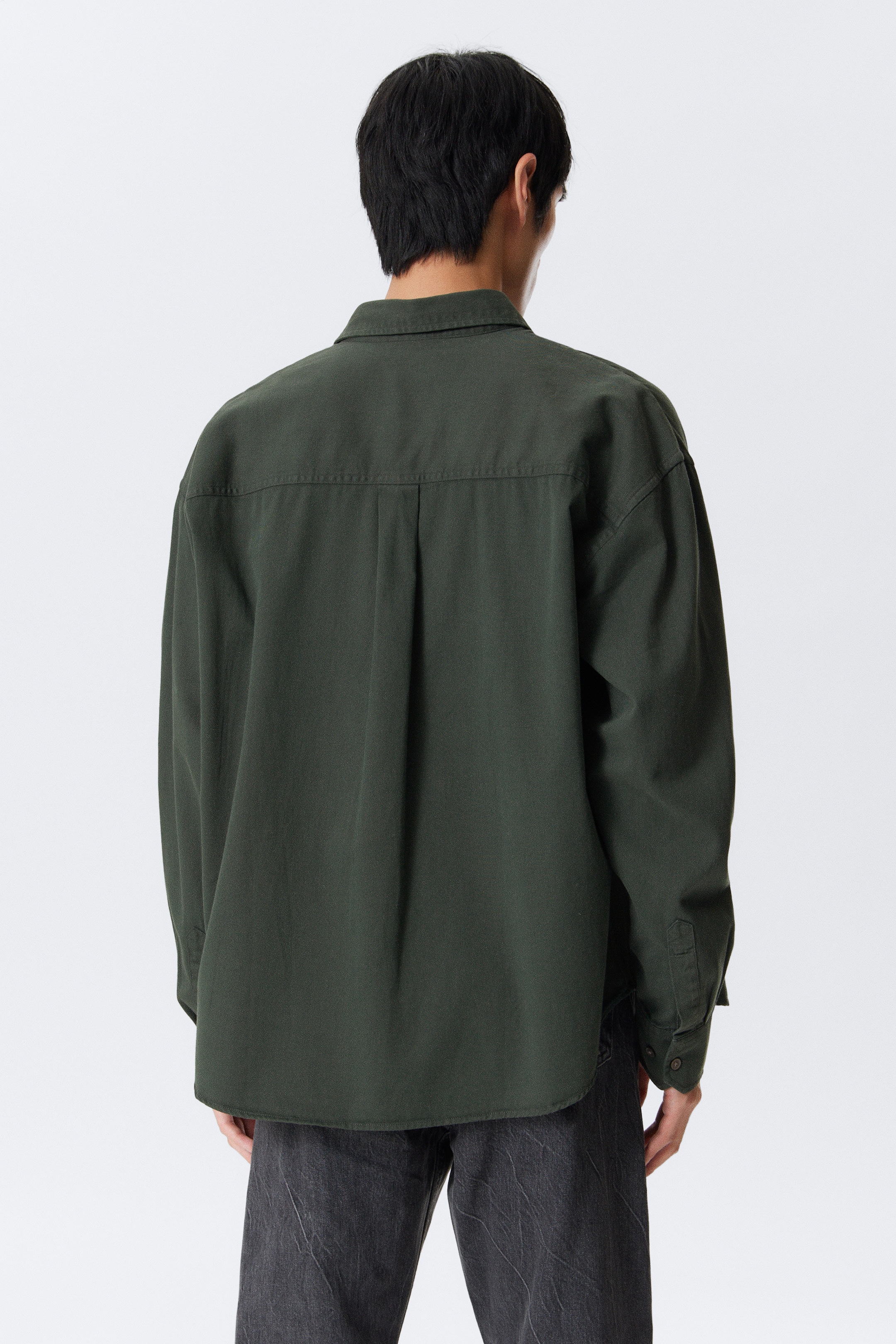 Se større billede: En mørkegrøn oversized skjorte ses bagfra, med en lige rygbærestykke og en central boksfold. Den er designet med lange ærmer, knappede manchetter og en afrundet kant.