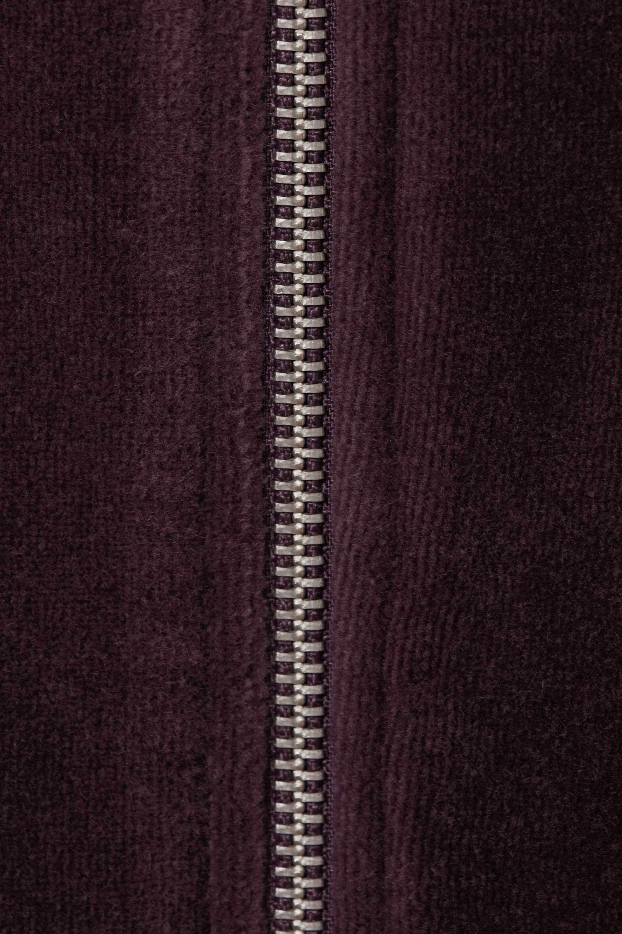 Agrandir l'image: Une veste en velours sombre présente une fermeture éclair de couleur argentée proéminente qui descend verticalement au centre, mettant en valeur la texture douce du tissu.
