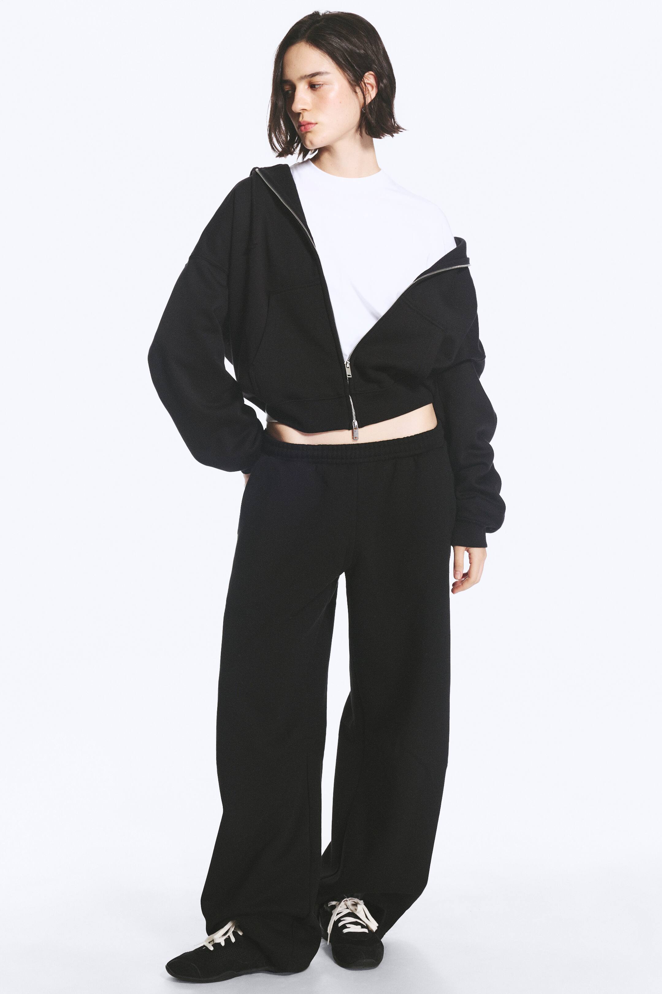 Agrandir l'image: Un pantalon de jogging noir à coupe ample, doté d’une taille élastiquée et d’une jambe arrondie de type barrel, est porté par le modèle. Le pantalon est associé à un hoodie court noir zippé assorti, à un t-shirt blanc et à des sneakers noires.