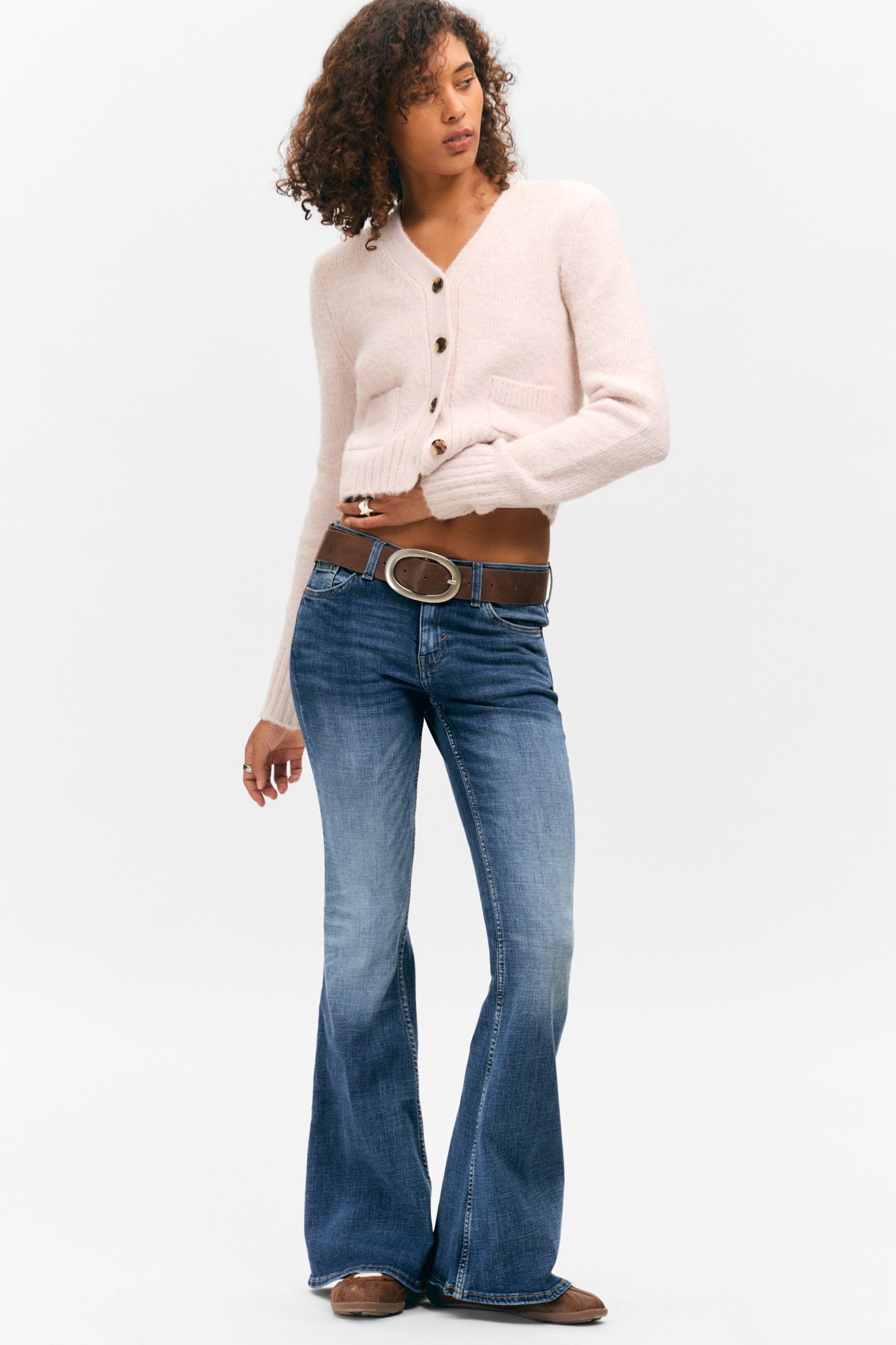 Ingrandisci l'immagine: Un cardigan corto in peluche di colore rosa chiaro presenta uno scollo a V, tre bottoni marroni, due tasche applicate anteriori e polsini e orlo a coste. Jeans a zampa blu mostrano un lavaggio sbiadito, aderendo lungo la coscia e svasando sotto il ginocchio, e sono abbinati a una cintura marrone con una fibbia ovale argentata.