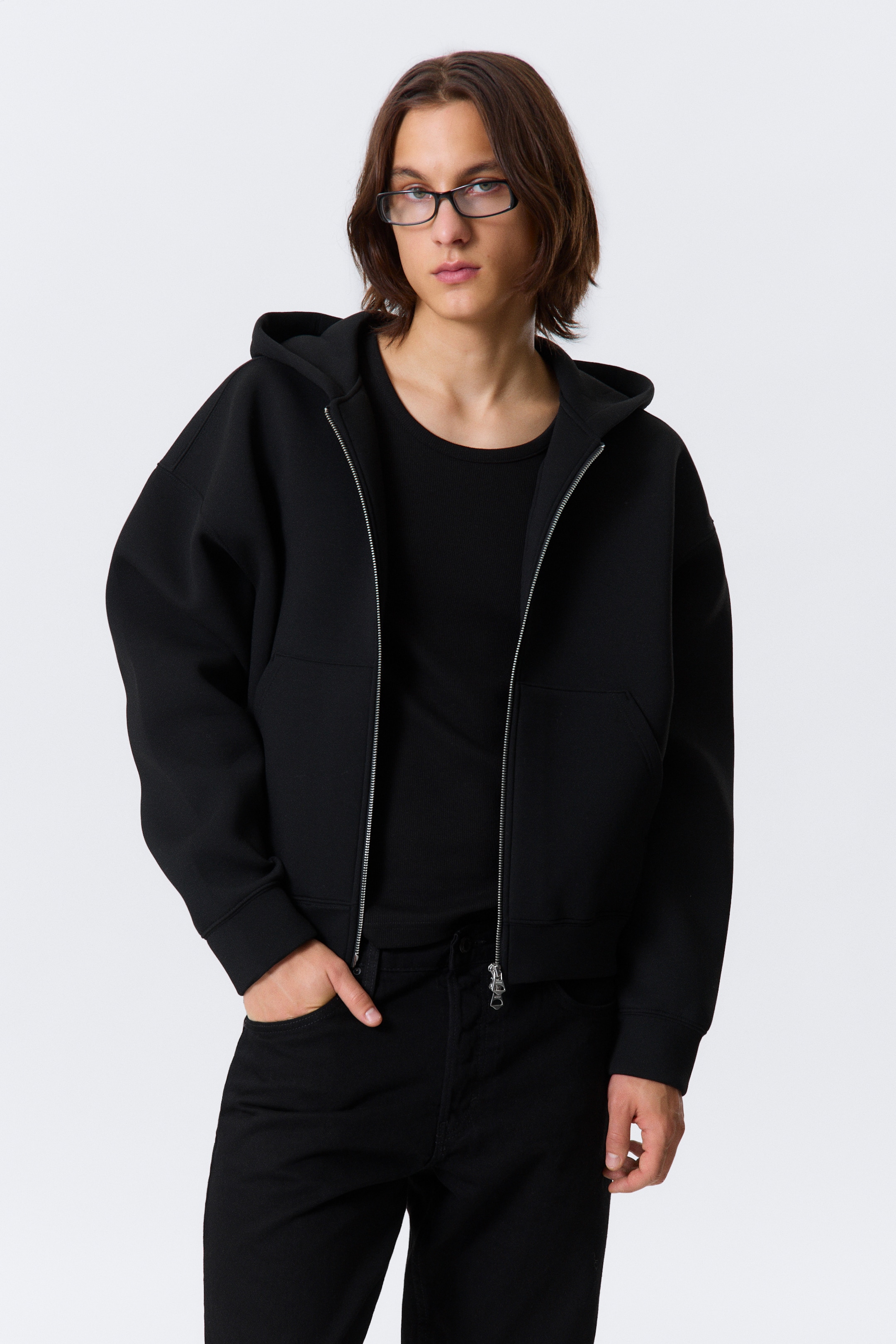 View larger image: A black full-zip hooded jacket is being presented, featuring a structured, cropped design with dropped shoulders and a silver-tone front zipper. It is being worn over a black ribbed crew-neck top.