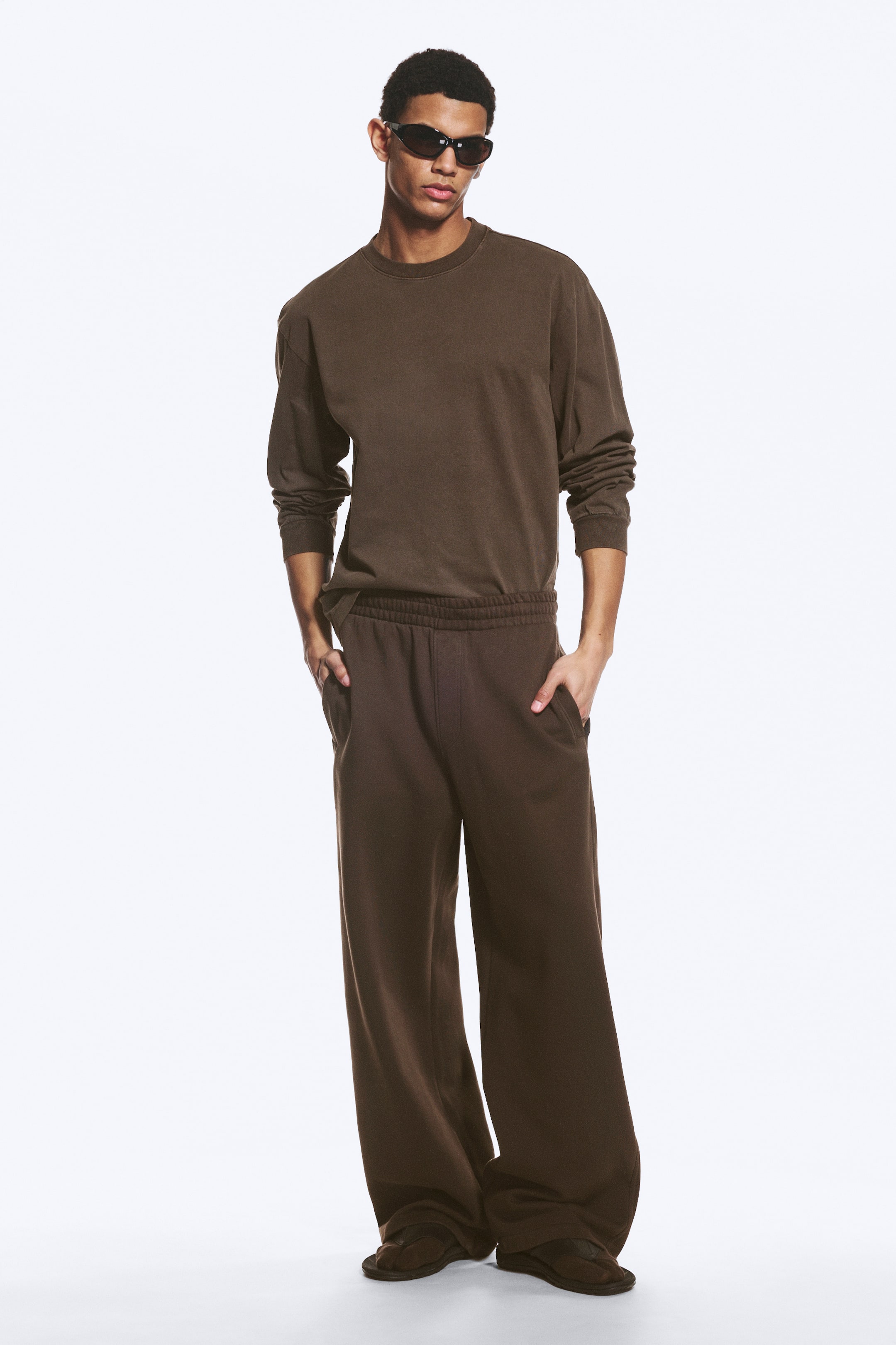 Grotere afbeelding bekijken: Een gecoördineerde set bruine loungewear wordt gepresenteerd vanuit een frontale, licht schuine hoek. De set bestaat uit een longsleeve crewneck t-shirt met een ontspannen pasvorm en wijde sweatpants met een elastische tailleband en zijzakken.