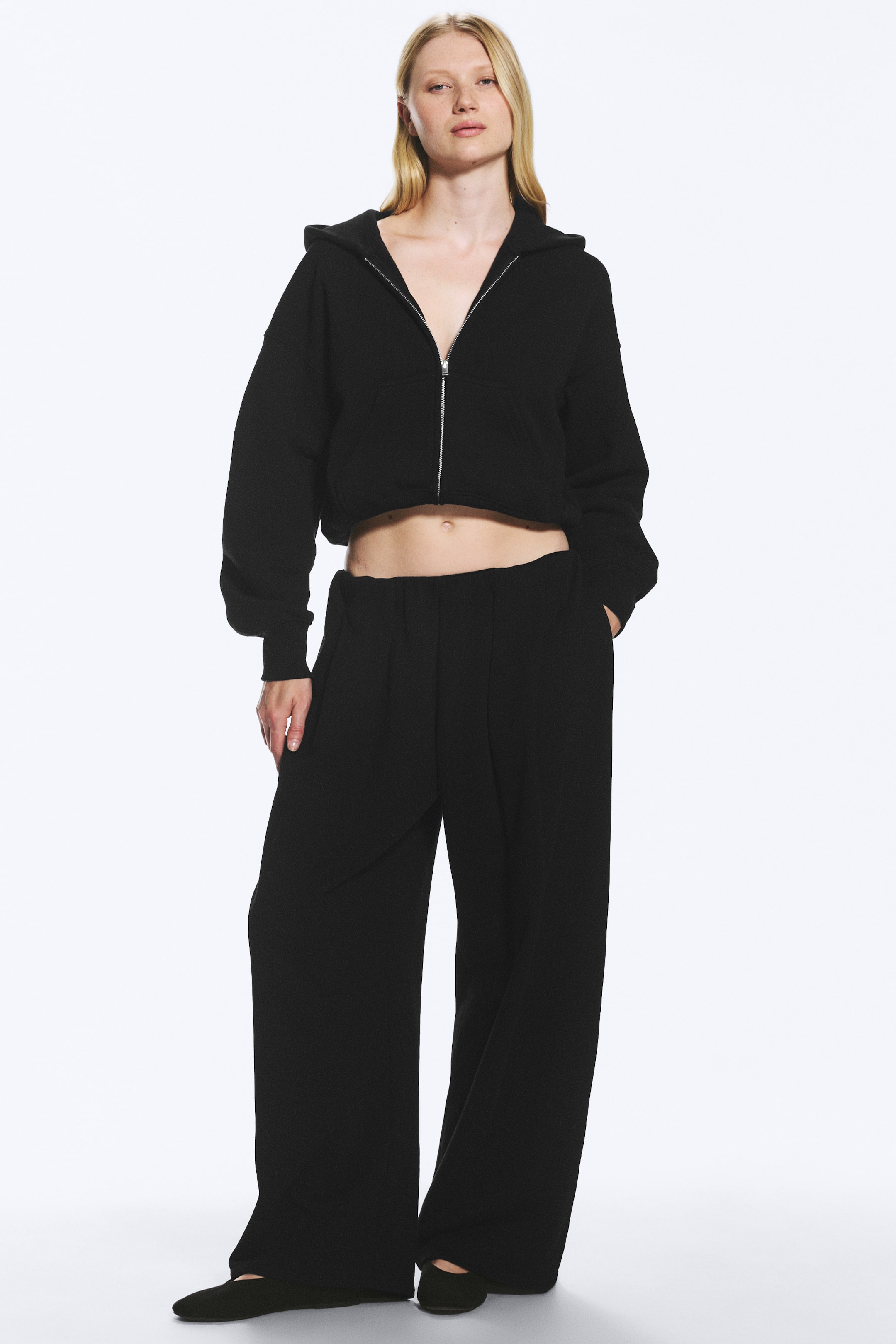 Agrandir l'image: Le mannequin porte un pantalon drapé en jersey. Ce pantalon noir présente une silhouette à jambes larges, une taille élastiquée et une couture apparente à l’avant sur chaque jambe. Le mannequin est debout, une main dans la poche du pantalon.