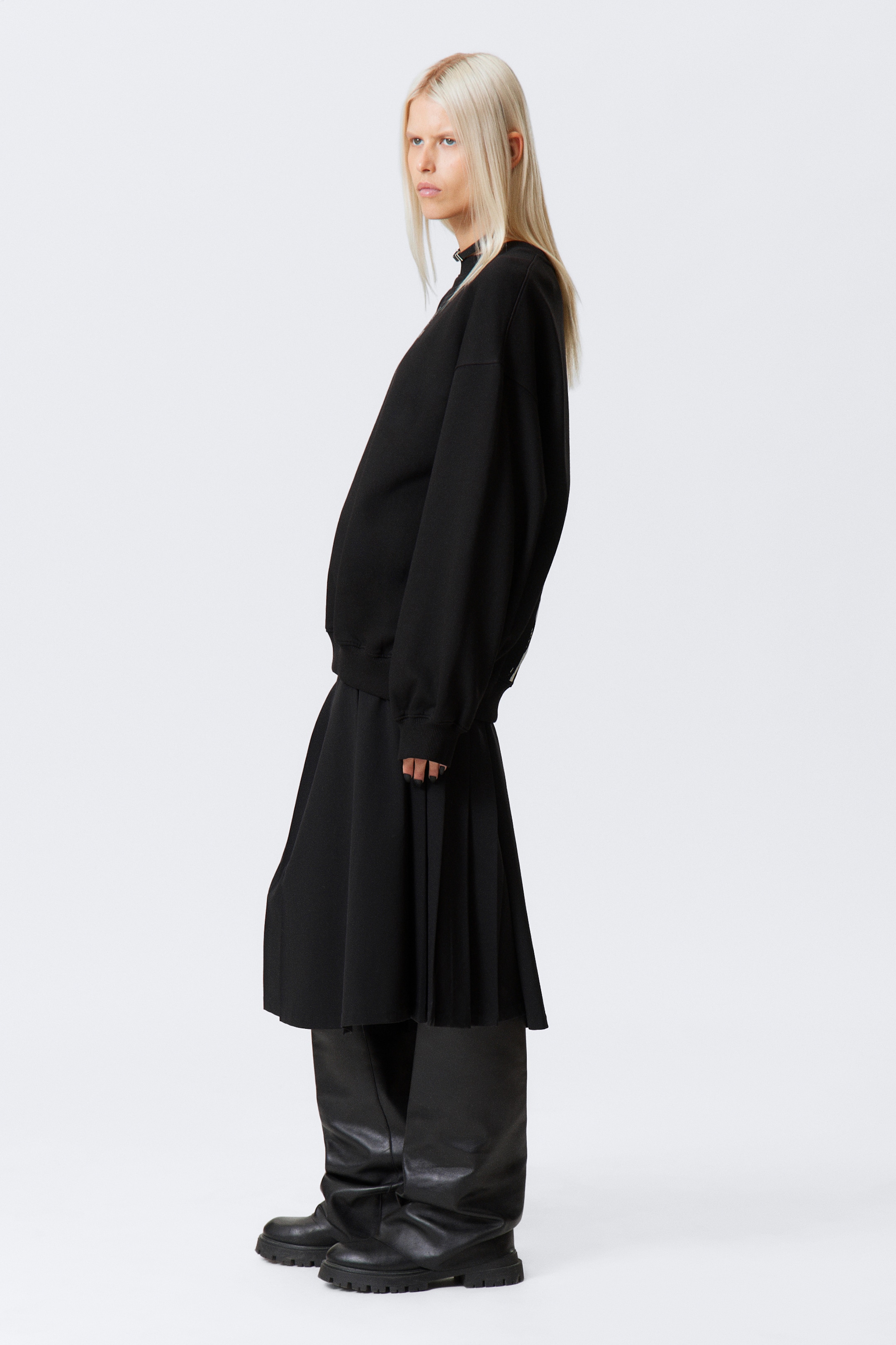 Visualizza immagine più grande: Una felpa nera oversize con girocollo e polsini a coste viene indossata, stratificata su una gonna midi nera plissettata. La gonna è stilizzata sopra pantaloni neri a gamba larga che terminano in stivali con suola a carrarmato, con l'intero ensemble presentato da un profilo laterale per enfatizzare la silhouette stratificata.