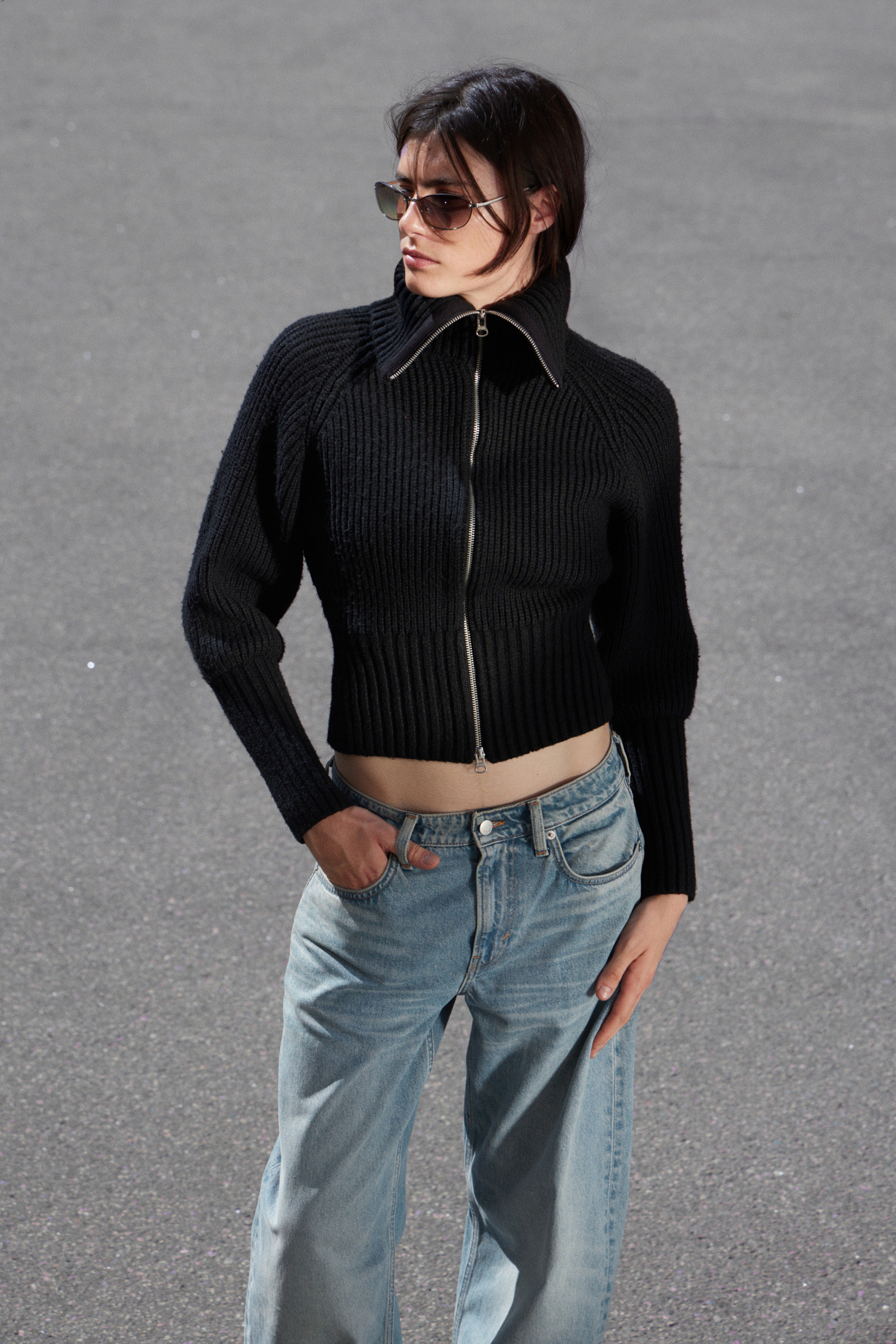 Grösseres Bild ansehen: Ein schwarzes geripptes Strick-Cropped-Top wird getragen, das einen breiten Stehkragen und einen silberfarbenen Reißverschluss in voller Länge aufweist. Das Top zeigt seinen eng anliegenden Saum und voluminöse lange Ärmel und wird mit hell gewaschenen Jeans gestylt.