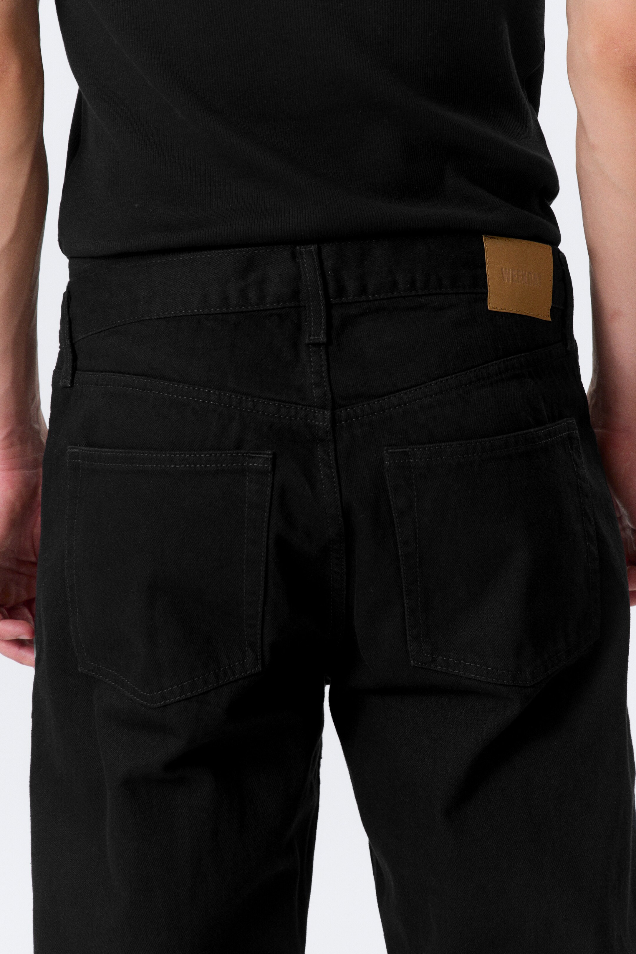 View larger image: Black jeans are being viewed from the back, featuring two rectangular patch pockets and subtle dark gray stitching. A light brown brand patch is visible on the right side of the waistband.