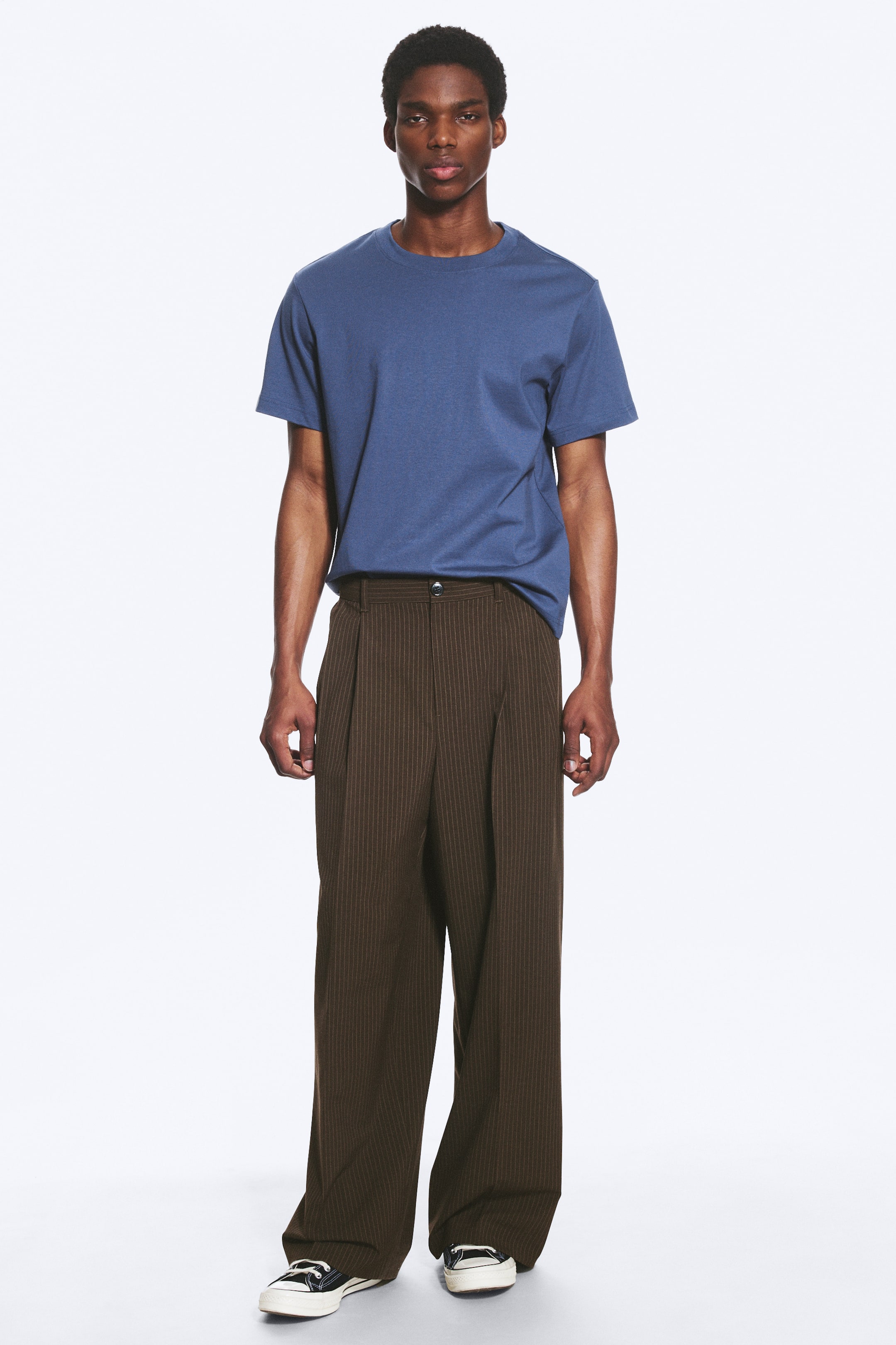Visualizza immagine più grande: I pantaloni del completo in twill a righe marroni sono indossati dal modello. Questi pantaloni larghi e dalla vestibilità ampia presentano pieghe frontali, tasche laterali inclinate visibili e pieghe stirate, e sono abbinati a una t-shirt blu e sneakers nere.