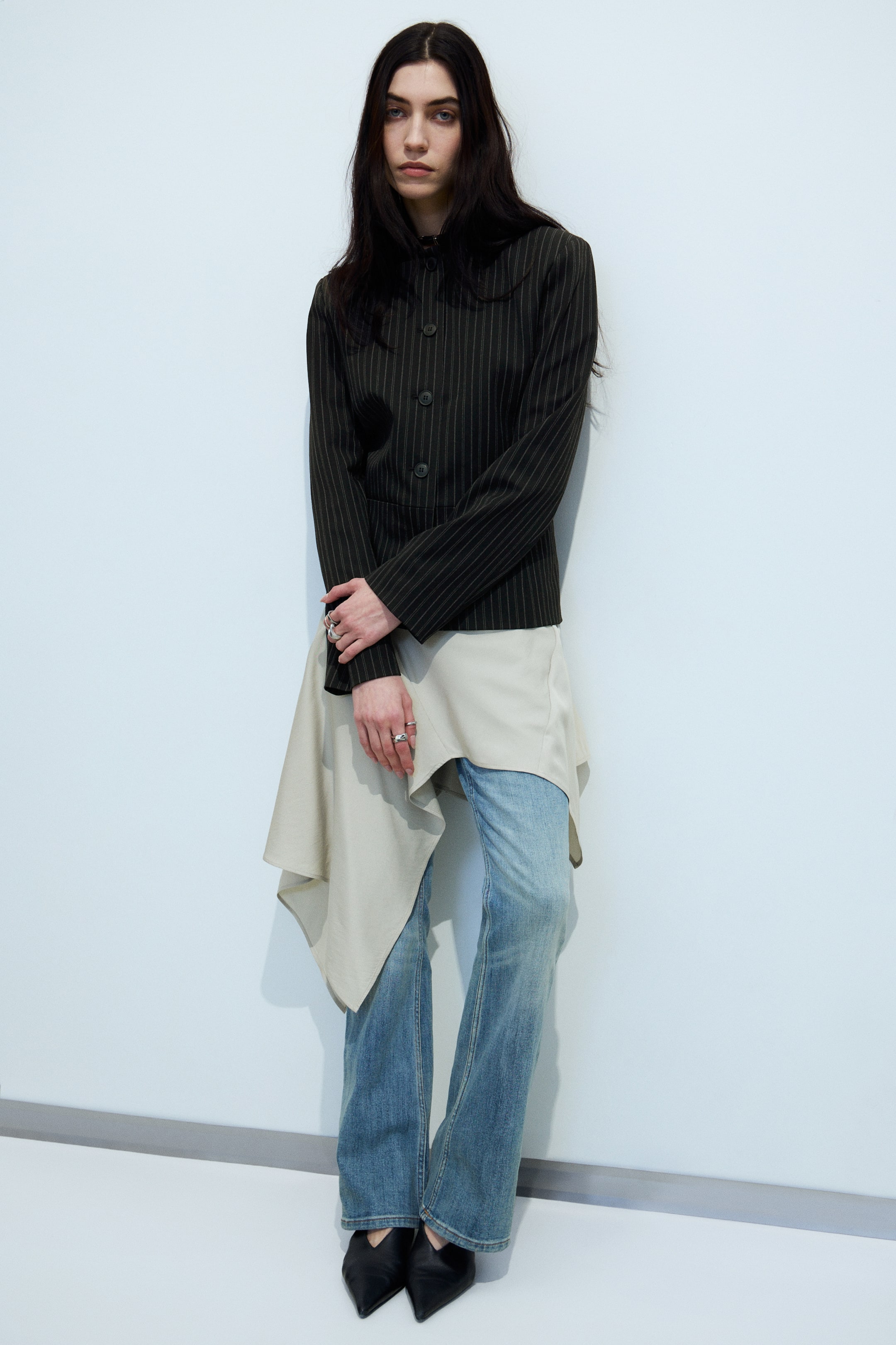 Visualizza immagine più grande: Una giacca a righe scure con colletto arrotondato e chiusura frontale a tre bottoni è sovrapposta a un top asimmetrico beige chiaro, che si estende sotto l'orlo della giacca. Jeans a gamba dritta azzurri completano i capi superiori, e scarpe basse a punta nera completano l'insieme, il tutto presentato frontalmente.