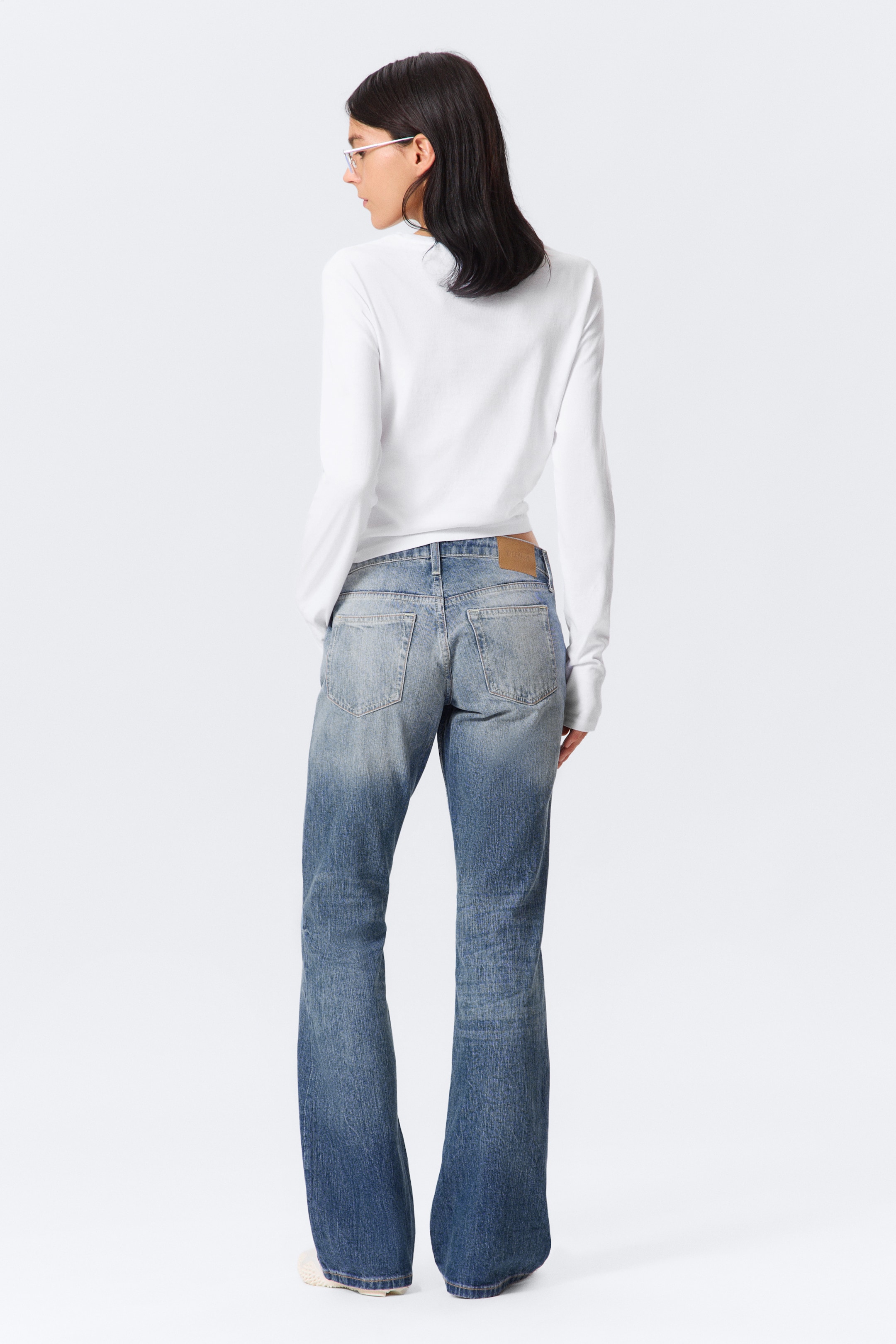 Grotere afbeelding bekijken: Blauwe, distressed jeans worden gepresenteerd vanuit een achterste driekwart hoek, met een vervaagde wassing die lichter wordt naar boven en donkerder naar de uitlopende zoom. De jeans met een relaxte pasvorm hebben zichtbare achterzakken en een lichtbruin label op de tailleband, en worden gedragen met een effen wit langemouwenshirt.