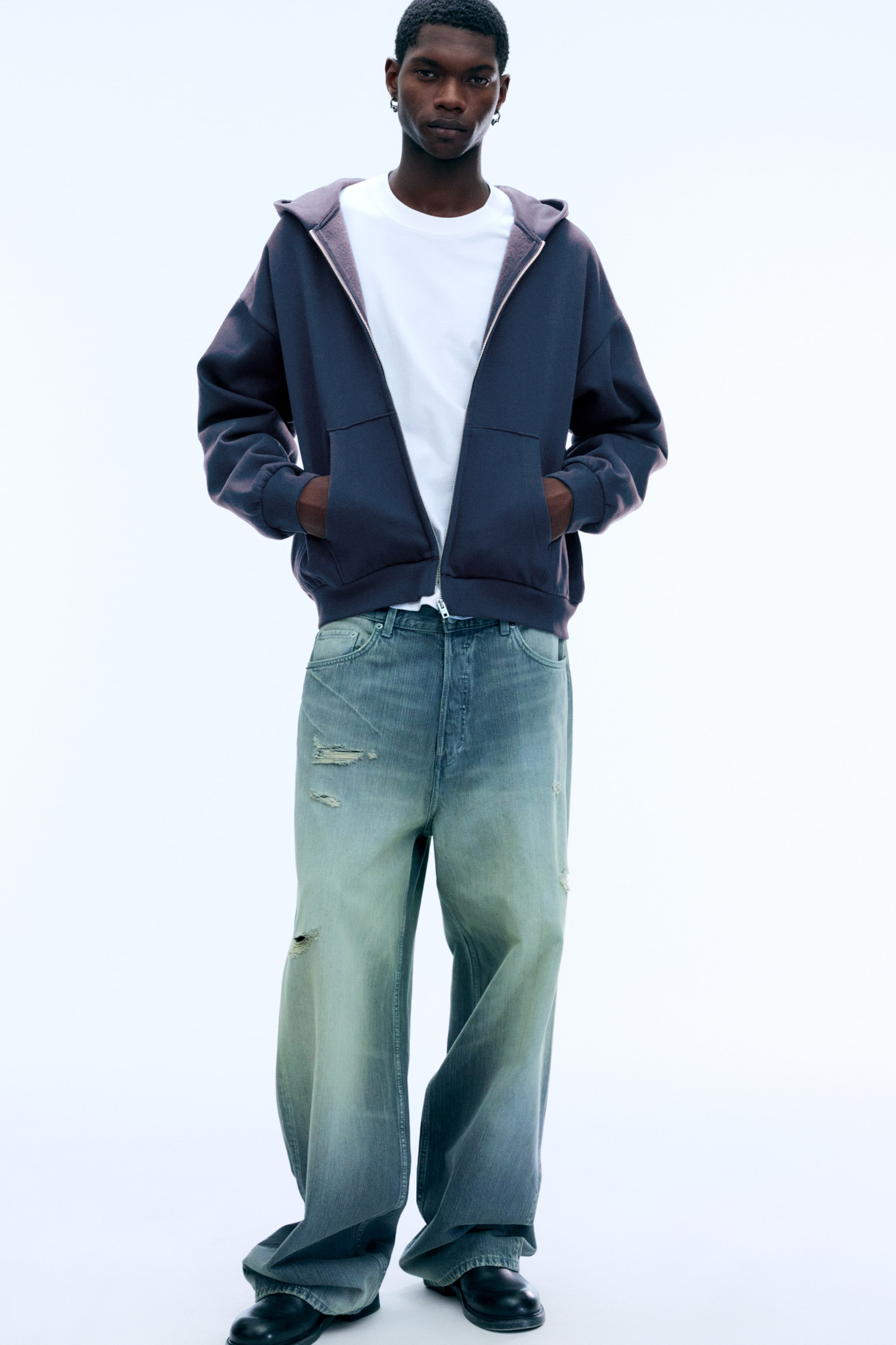 Zobacz większy obraz: Luźna, ciemnoszara bluza z kapturem zapinana na zamek, z obniżoną linią ramion i marszczonym dołem, jest noszona przez modela, który stoi z dłońmi w przednich kieszeniach. Bluza jest założona na biały T-shirt i zestawiona z szerokimi, przetartymi jeansami z denimu.