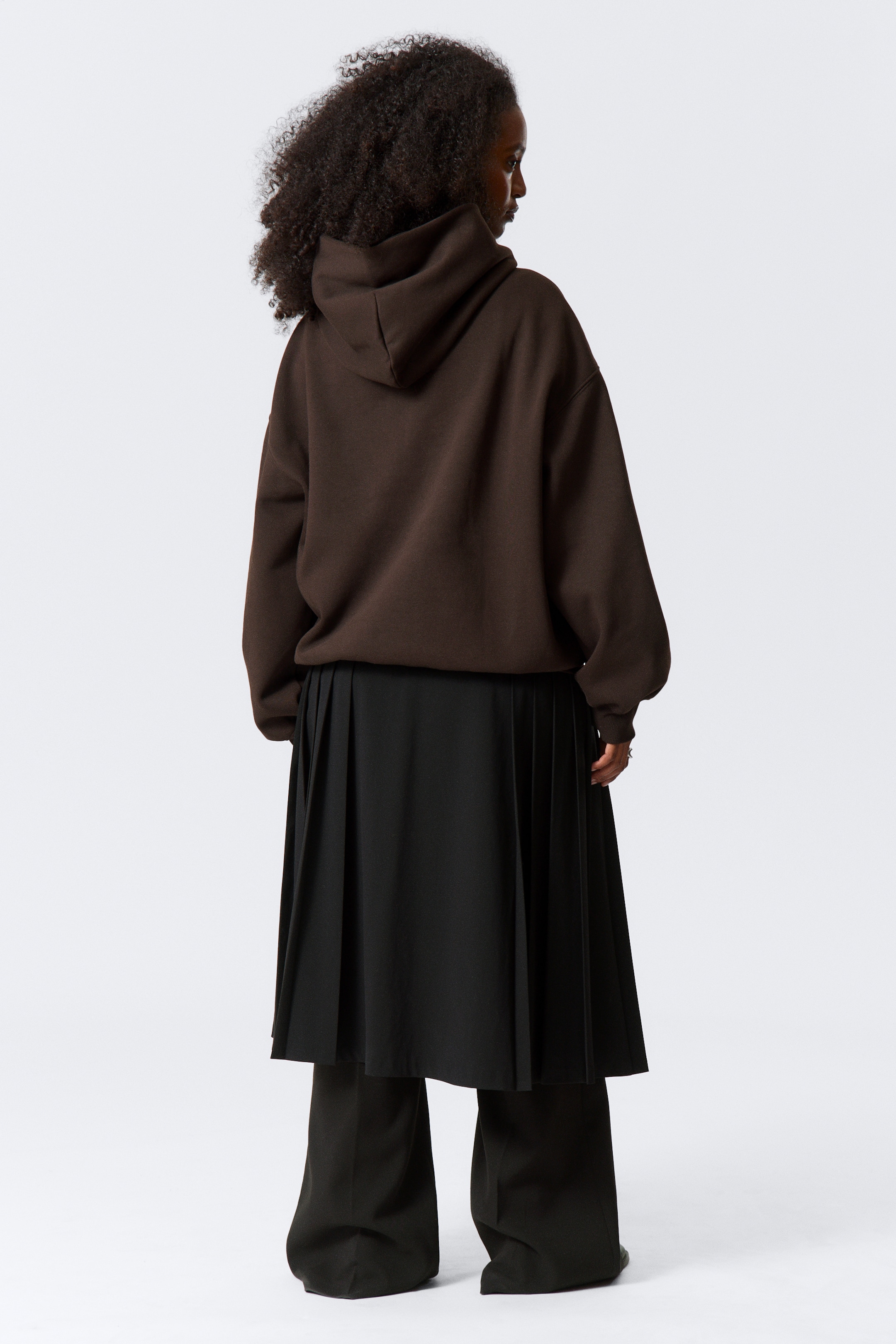 Visa större bild: En mörkbrun oversized hoodie bärs, med en synlig huva och långa ärmar med elastiska muddar. Den är lagrad över en svart midilång plisserad kjol, som sedan är lagrad över mörka vida byxor. Alla plagg visas bakifrån.