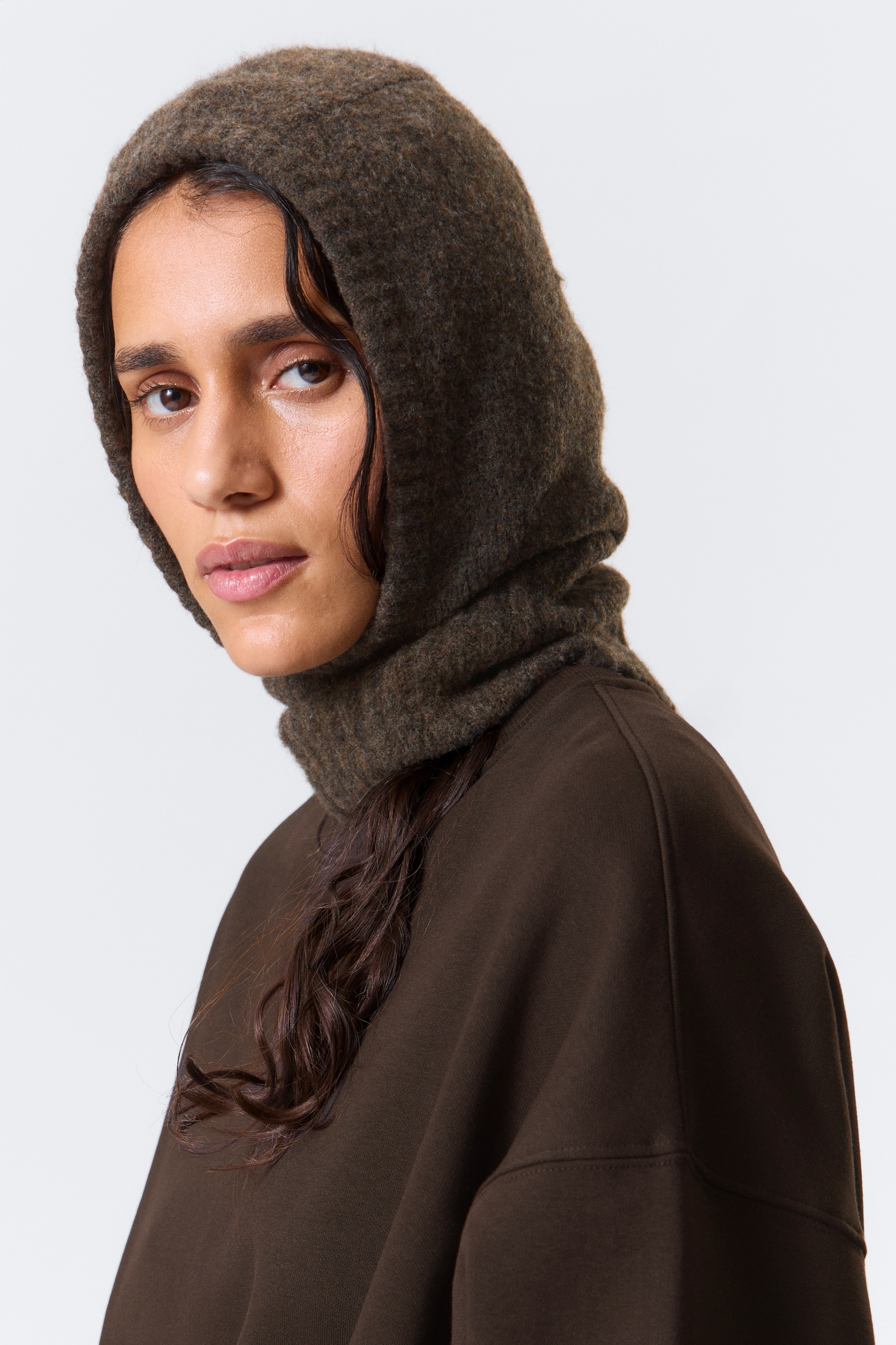 View larger image: A dark olive green, fuzzy knit balaclava-style hood is covering the head and neck, extending over the shoulders. The hood section is fitting closely around the face, while the neck section is loosely draped over a dark brown long-sleeved top.