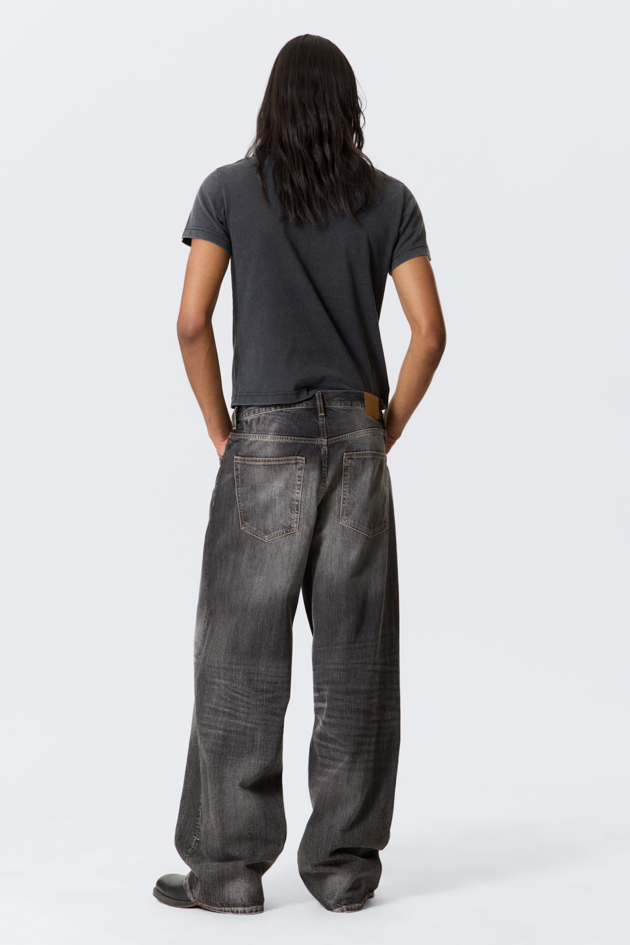 Ingrandisci l'immagine: Un paio di jeans in denim nero slavato e ampio viene mostrato da dietro, caratterizzato da una finitura sbiadita, tasche posteriori visibili e una silhouette a gamba larga che si accumula alle caviglie. Una t-shirt a maniche corte grigio scuro viene indossata con i jeans.