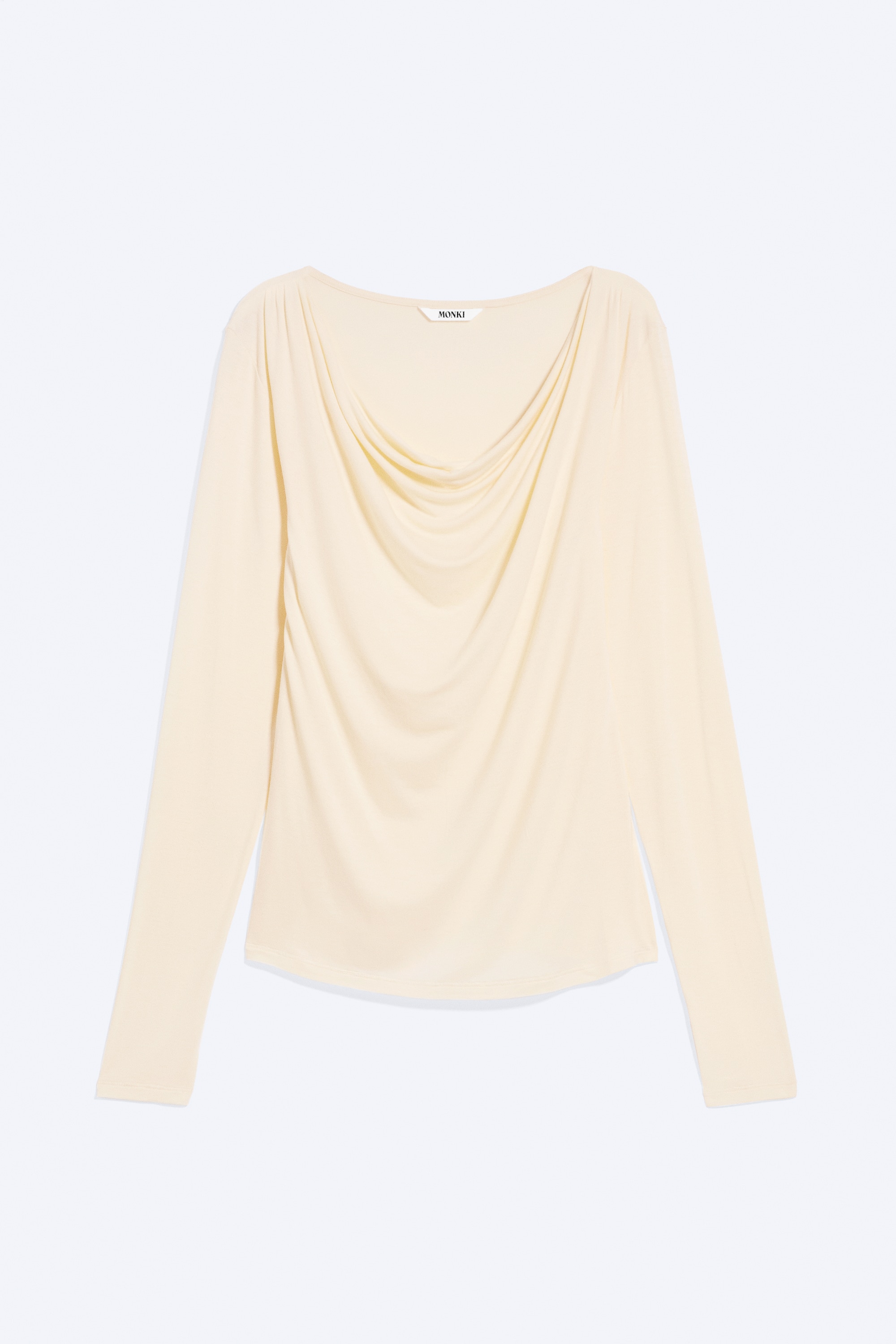 La modella indossa un top slim fit a maniche lunghe di colore giallo chiaro, caratterizzato da una scollatura asimmetrica drappeggiata tirata giù su una spalla e da arricciature lungo il corpo. Il top è abbinato a jeans blu chiaro lavati, lasciando intravedere una sottile spallina nera sulla spalla della modella.