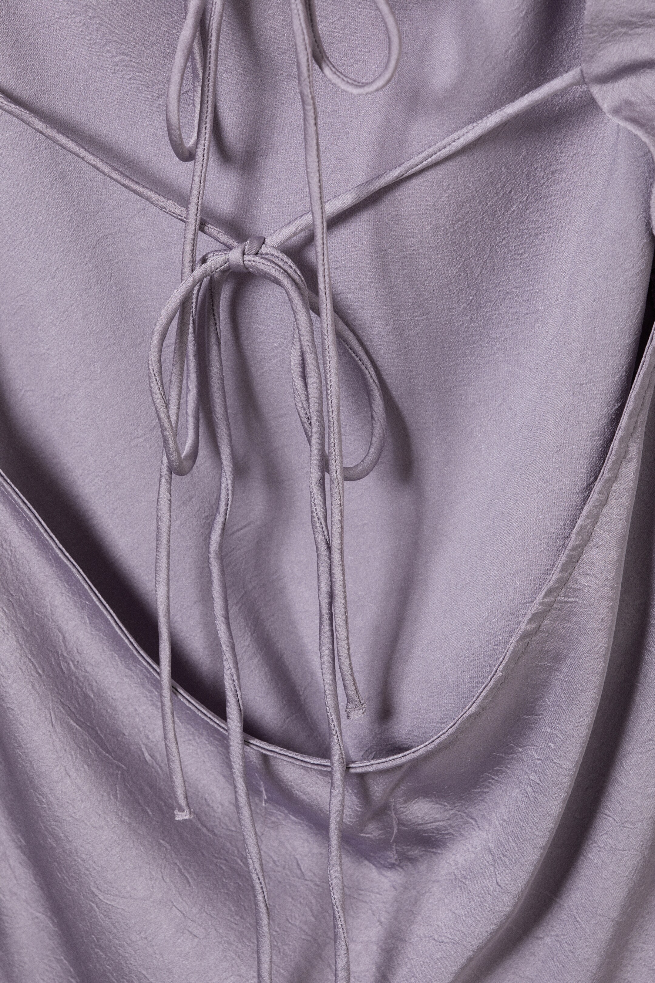 View larger image: The back of a lavender, textured satin-like garment is being shown, featuring thin straps crisscrossing and tying in a knot, above a deep, draped open back.