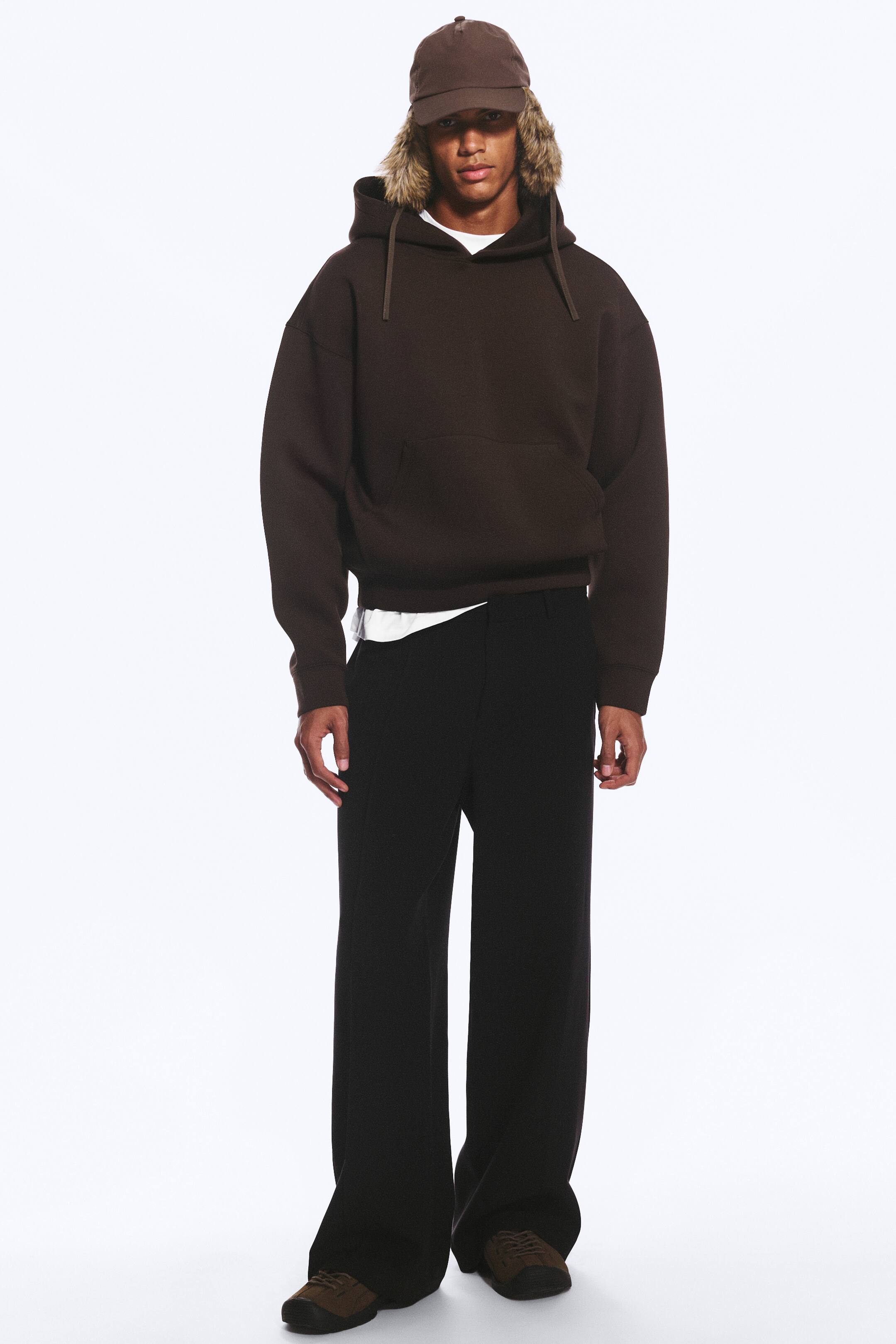 Ver imagem maior: Calças de fato pretas, de corte descontraído e perna direita, estão a ser usadas pela modelo. As calças são combinadas com um hoodie castanho-escuro, uma t-shirt branca visível na cintura e sapatos castanhos.