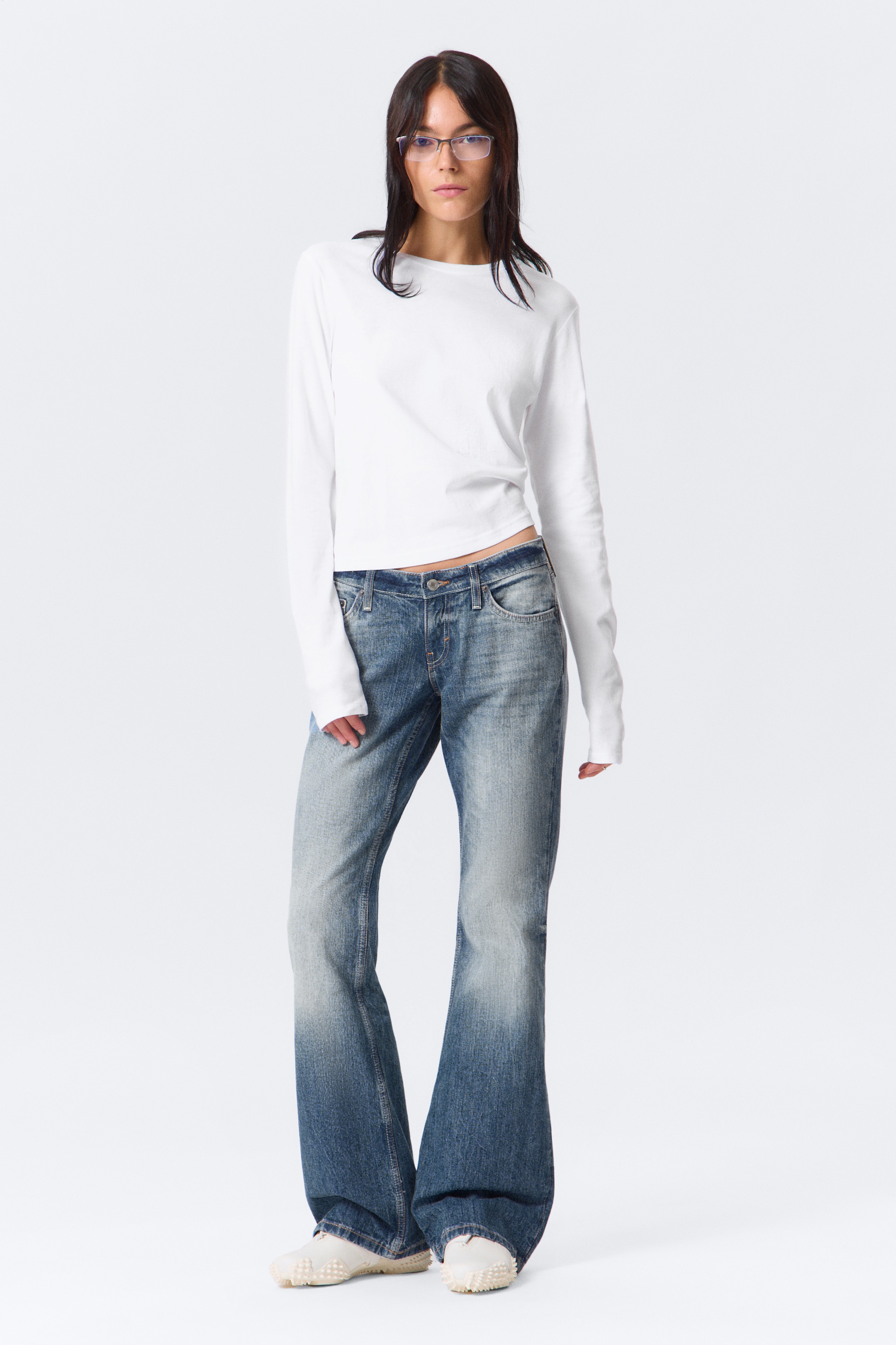 Grotere afbeelding bekijken: Een wit top met lange mouwen met een ronde hals en een iets kortere zoom wordt gepresenteerd. Het is gestyled met blauwe denim jeans, die een vervaagde wassing op de dijen tonen en een uitlopende snit aan de zoom hebben.