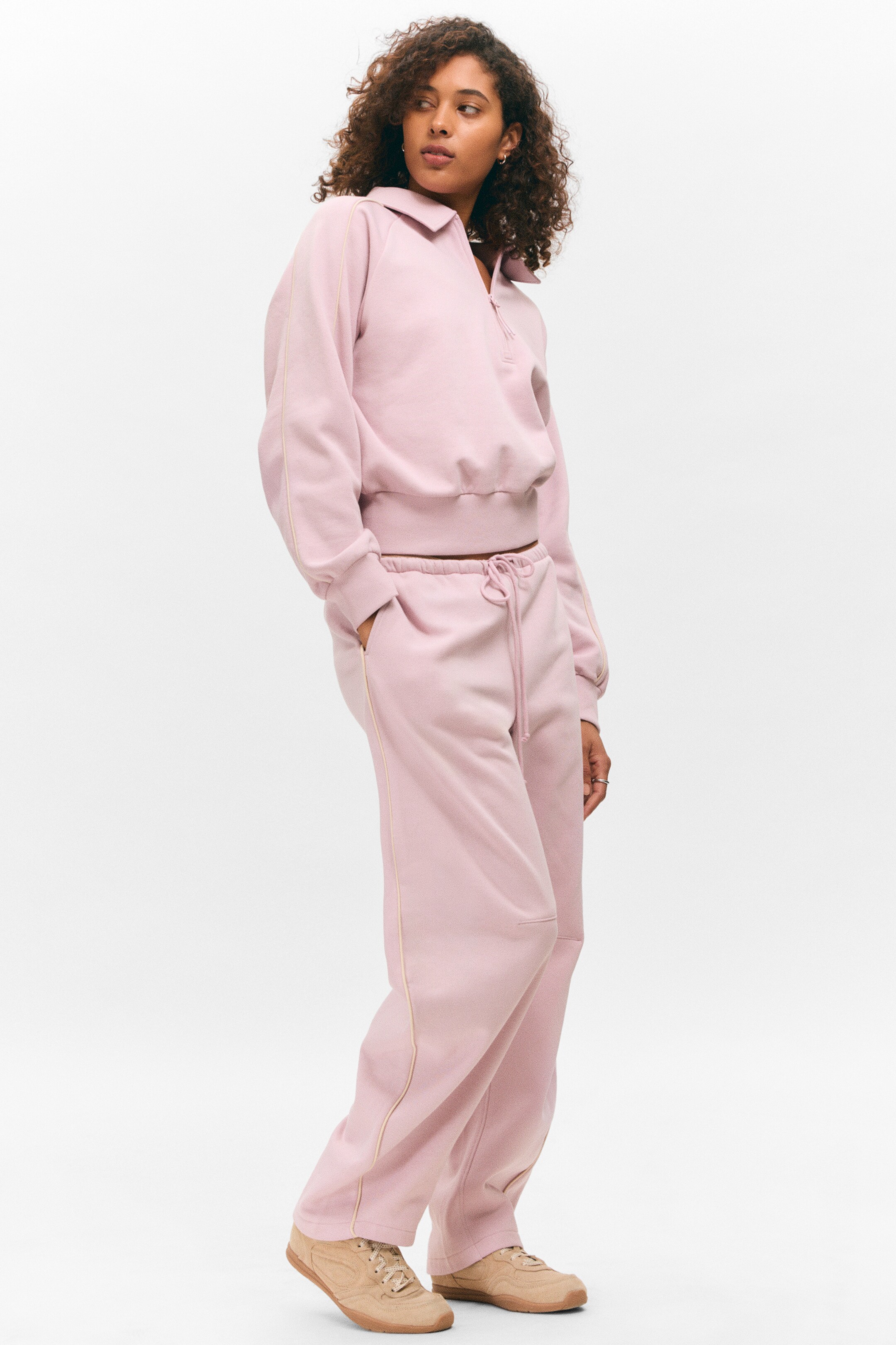 Agrandir l'image: Un survêtement rose est présenté, orienté légèrement vers la droite. Le sweat-shirt à col et à quart de zip présente un passepoil blanc le long de ses manches raglan, des poignets côtelés et un ourlet côtelé. Le pantalon de survêtement assorti, à coupe décontractée, arbore une taille à cordon de serrage et un passepoil blanc le long de ses coutures latérales, tandis que des baskets à lacets marron clair complètent la tenue.