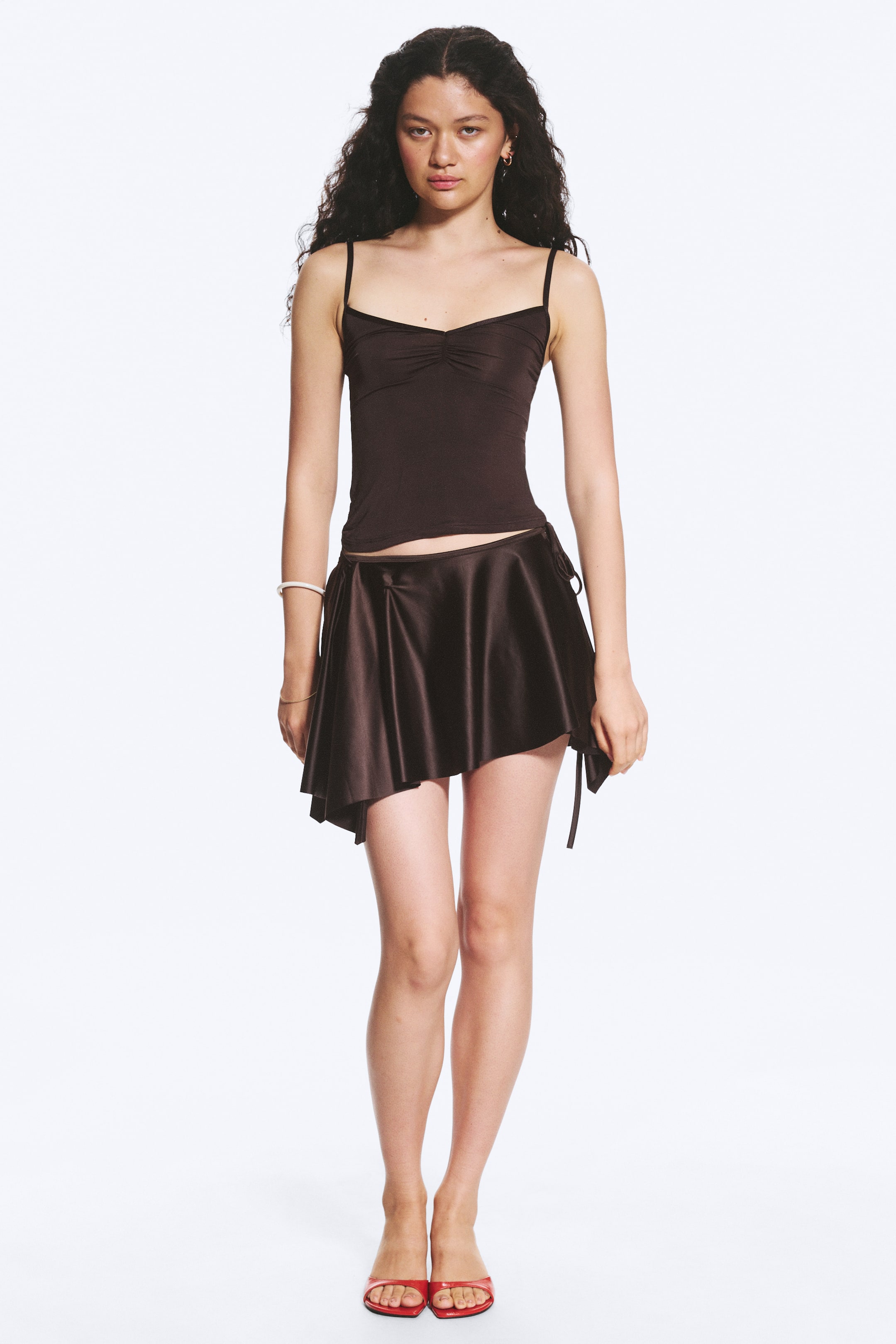 Zobacz większy obraz: Ciemnobrązowy top na ramiączkach charakteryzuje się dekoltem w kształcie serca z marszczeniem na środku oraz cienkimi ramiączkami typu spaghetti. Jest zestawiony z ciemnobrązową spódniczką mini o satynowym wyglądzie, z asymetrycznym brzegiem i bocznymi wiązaniami, które tworzą efekt drapowania.