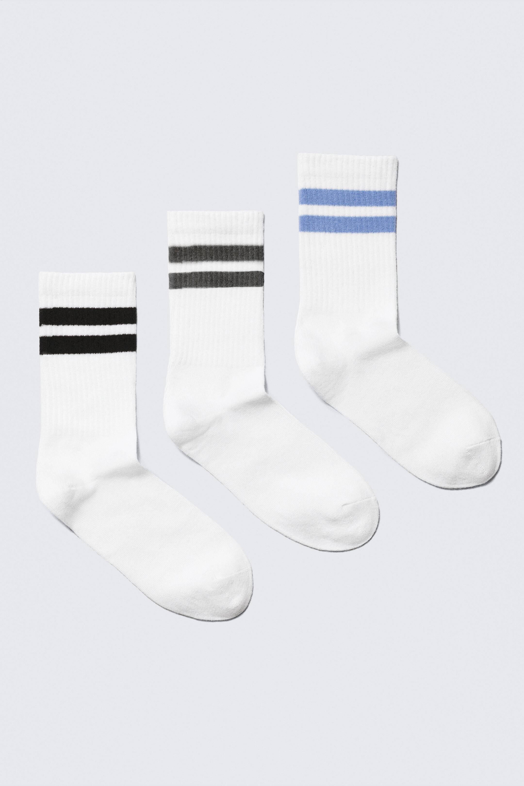Grotere afbeelding bekijken: Drie witte crew sokken liggen naast elkaar, elk met een geribbelde schacht en twee horizontale strepen. Eén sok heeft zwarte strepen, een andere heeft donkergrijze strepen, en de derde heeft lichtblauwe strepen.