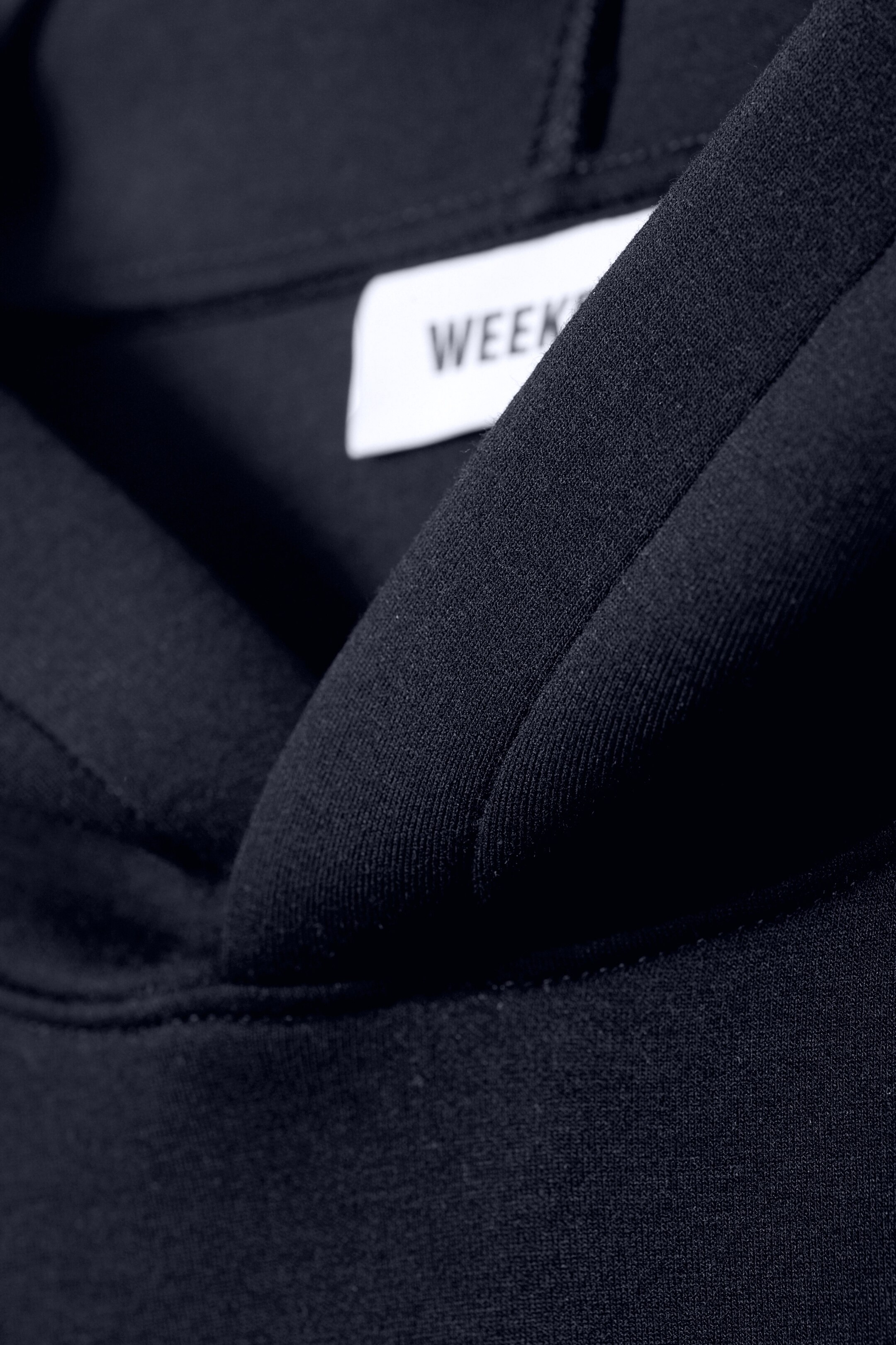 Größeres Bild anzeigen: Ein schwarzer Hoodie wird in einer Nahaufnahme aus einem schrägen Blickwinkel präsentiert, wobei das Innere seiner Kapuze und seines Halsausschnitts hervorgehoben wird. Die strukturierte, doppellagige Konstruktion der Kapuze ist sichtbar, zusammen mit einem weißen Markenschild, das teilweise „WEEK“ anzeigt und im hinteren Nacken positioniert ist.
