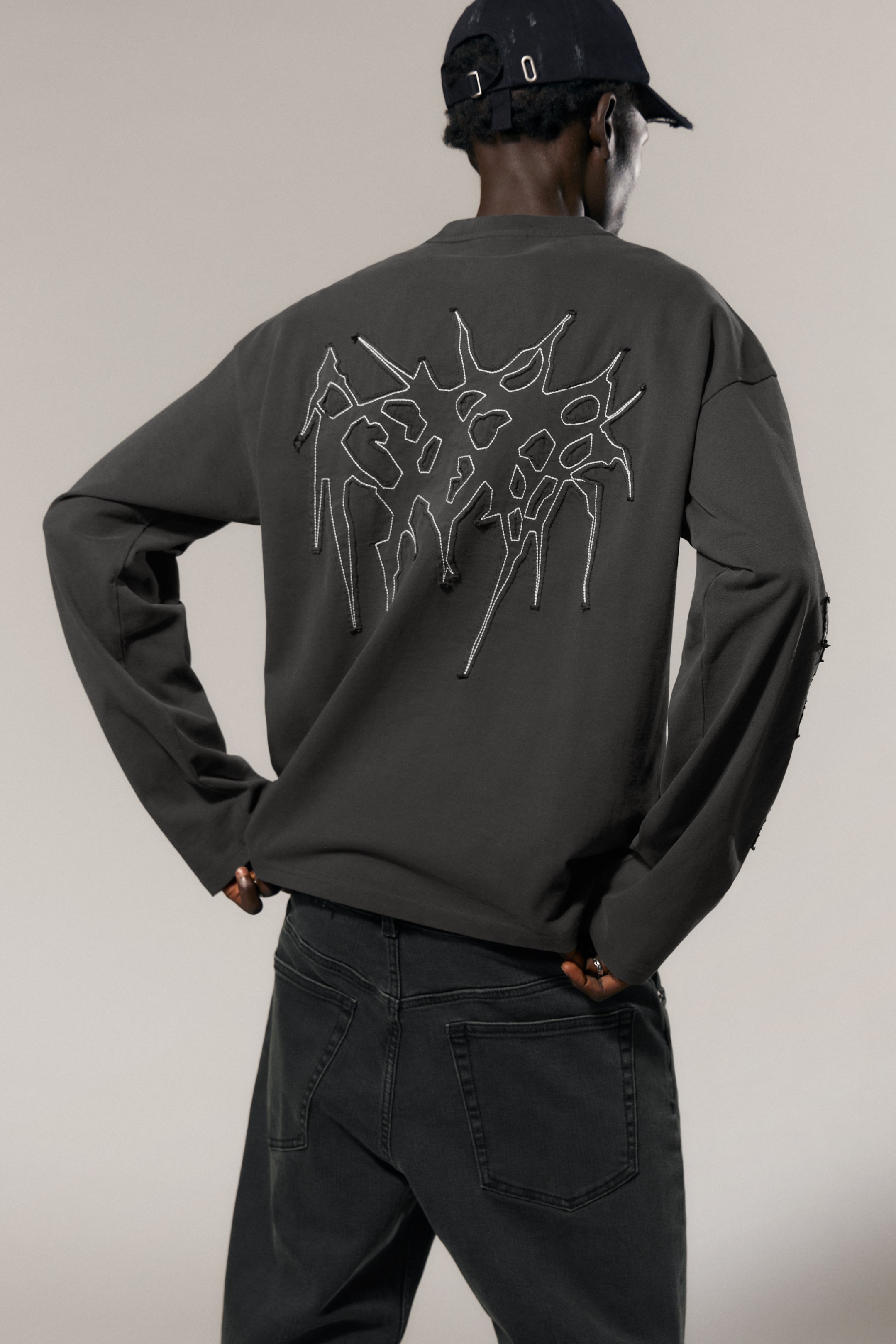 Größeres Bild ansehen: Ein dunkelgraues Langarmshirt mit Rundhalsausschnitt, von hinten präsentiert, zeigt ein großes weißes abstraktes Umrissdesign mit auffälligen Nähten und erhabenen, unregelmäßigen Formen auf dem Rücken. Der rechte Ärmel weist ebenfalls kleine, distressed Cut-Outs entlang der äußeren Naht auf.